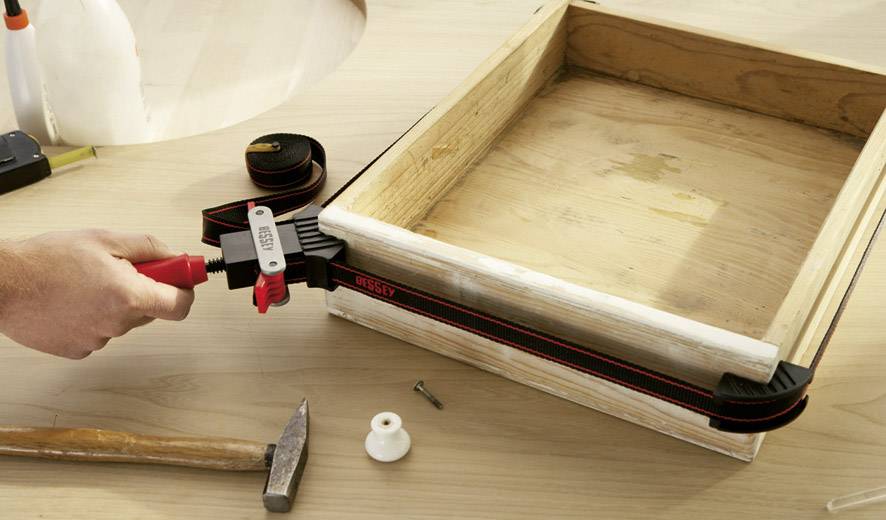 Eine Hand zieht mit einem Spannbandrahmen Kiste zusammen. Daneben liegen ein Hammer, eine Schraube und Holzstifte auf einem Tisch.