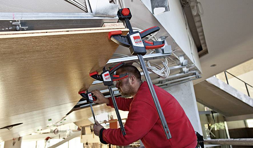Ein Handwerker befestigt eine Holzplatte an der Decke mit großen Zwingen in einem modernen Gebäude.