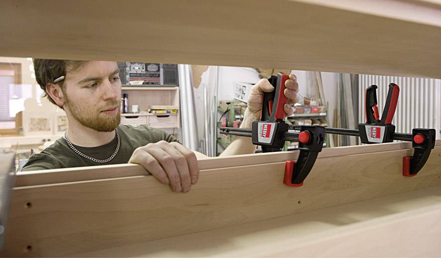 Ein Mann benutzt zwei Klemmen, um Holzbretter in einer Werkstatt zu fixieren. Im Hintergrund sind Werkzeuge und Holzarbeiten sichtbar.