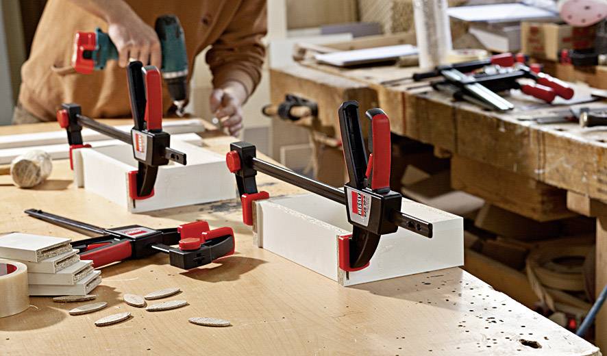 Person in Werkstatt fixiert Holz mit schwarzen und roten Schraubzwingen auf einer Arbeitsplatte, bereitet weiteres Werkstück vor.