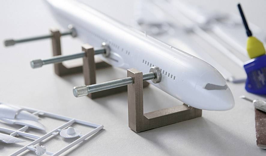 Ein Kunststoff-Modellflugzeug wird mit Schraubzwingen an Holzblöcken fixiert. Im Hintergrund sind Einzelteile und Modellbauzubehör zu sehen.