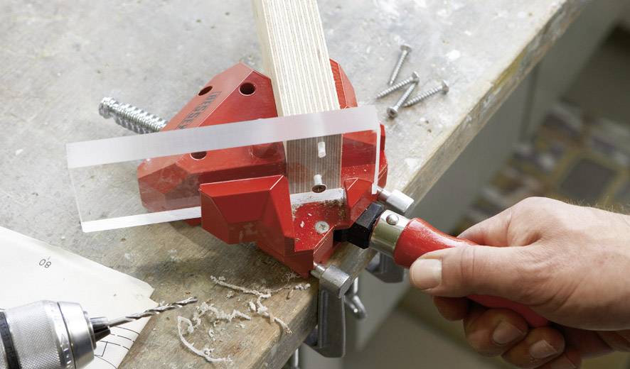 Eine Hand hält eine rote Gehrungssäge an einem Tisch und schneidet ein Holzstück. Schrauben und eine Bohrmaschine liegen daneben.
