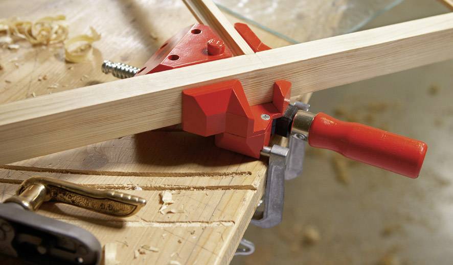 Ein Holzwerkstück wird mit einer roten Schraubzwinge auf einer Werkbank fixiert, umgeben von Holzspänen und Werkzeugen.