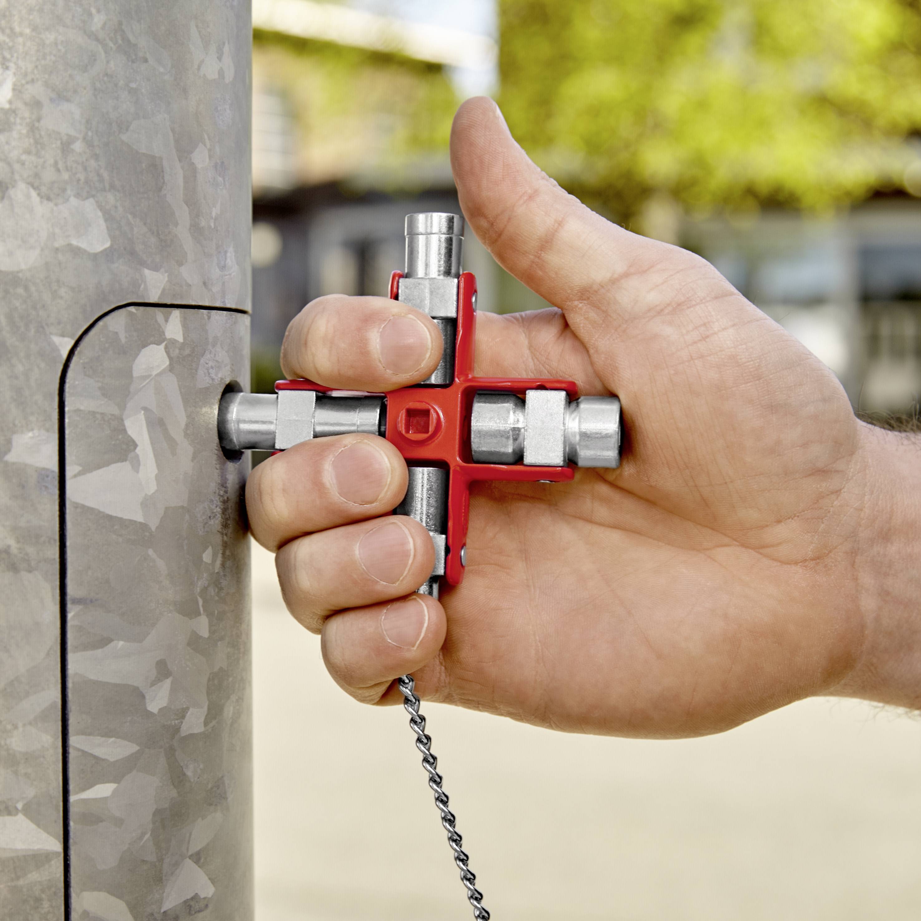 Hand hält roten Vierkantschlüssel mit mehreren Funktionen vor einem Metallpfosten. Hintergrund leicht unscharf, zeigt eine natürliche Umgebung.