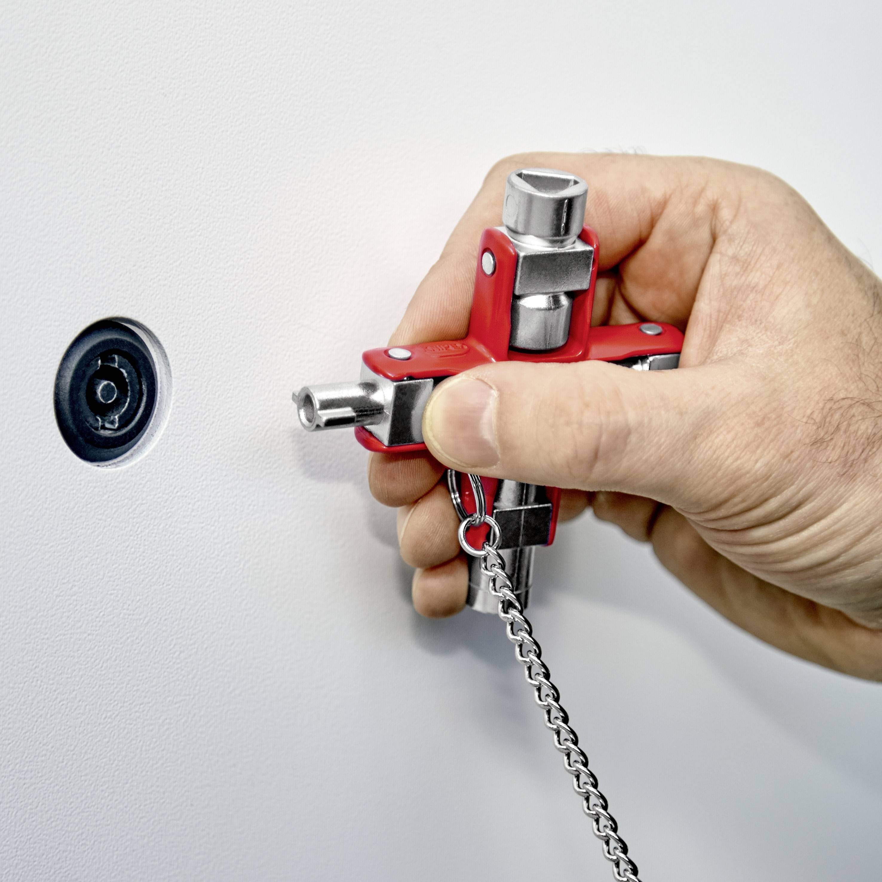 Eine Hand hält einen roten Vierkantschlüssel und versucht, das Schloss einer Tür zu öffnen. Der Schlüssel hängt an einer Metallkette.