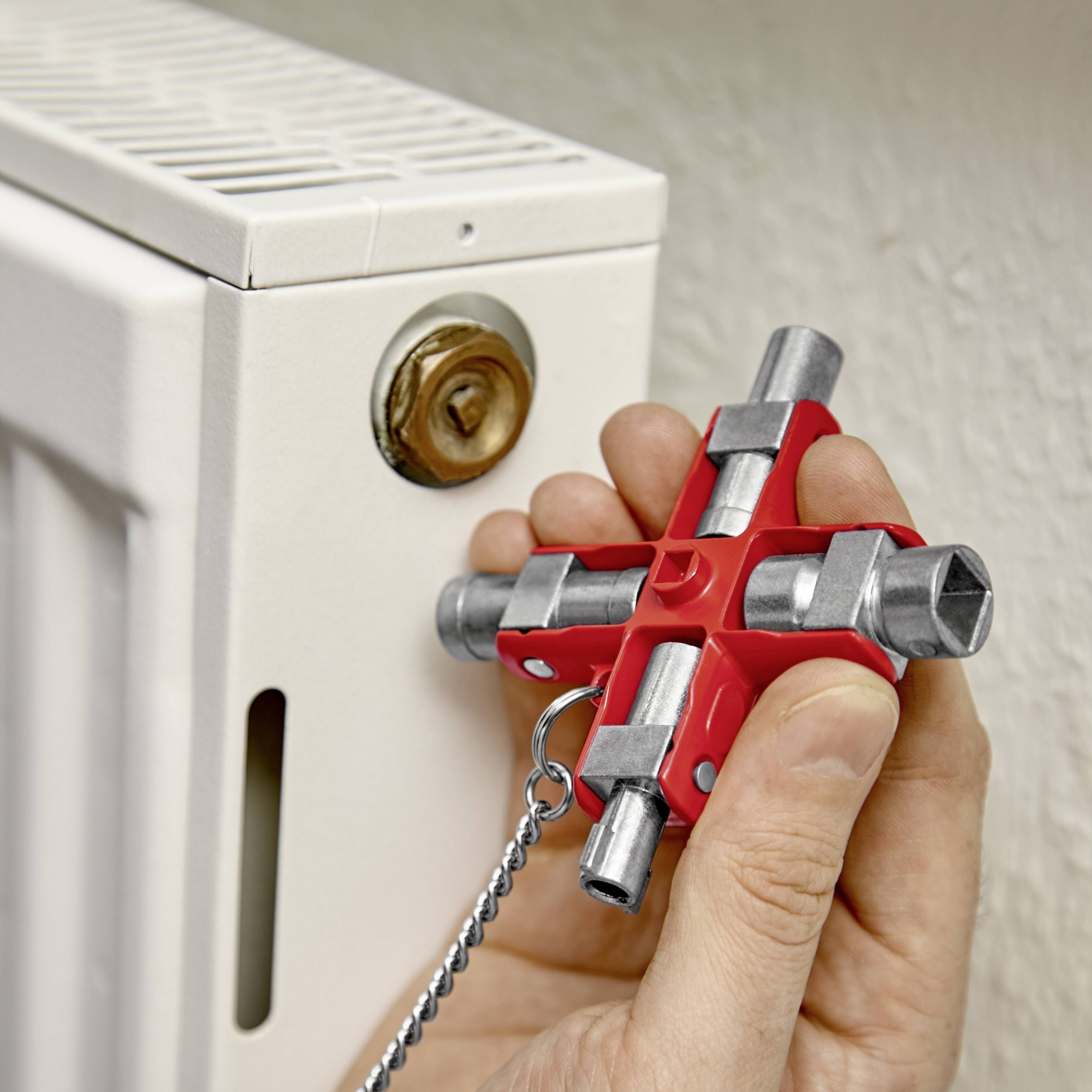 Eine Person hält einen Heizungsschlüssel und öffnet das Entlüftungsventil eines Heizkörpers. Der Schlüssel hat vier unterschiedliche Enden.
