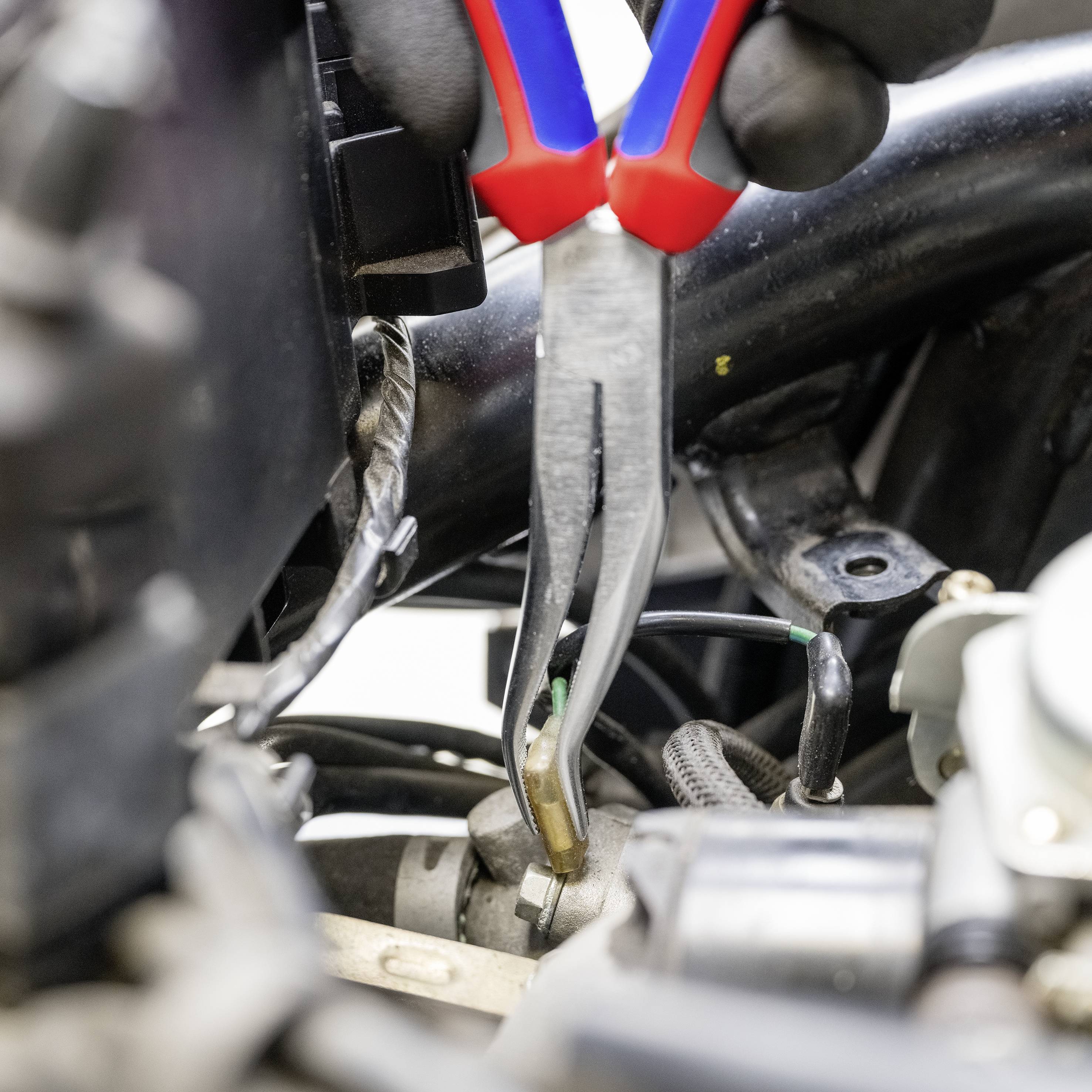 Ein Mechaniker verwendet eine Zange, um an einem elektrischen Kabel in einem Motorraum zu arbeiten. Kabel und Bauteile sind sichtbar.