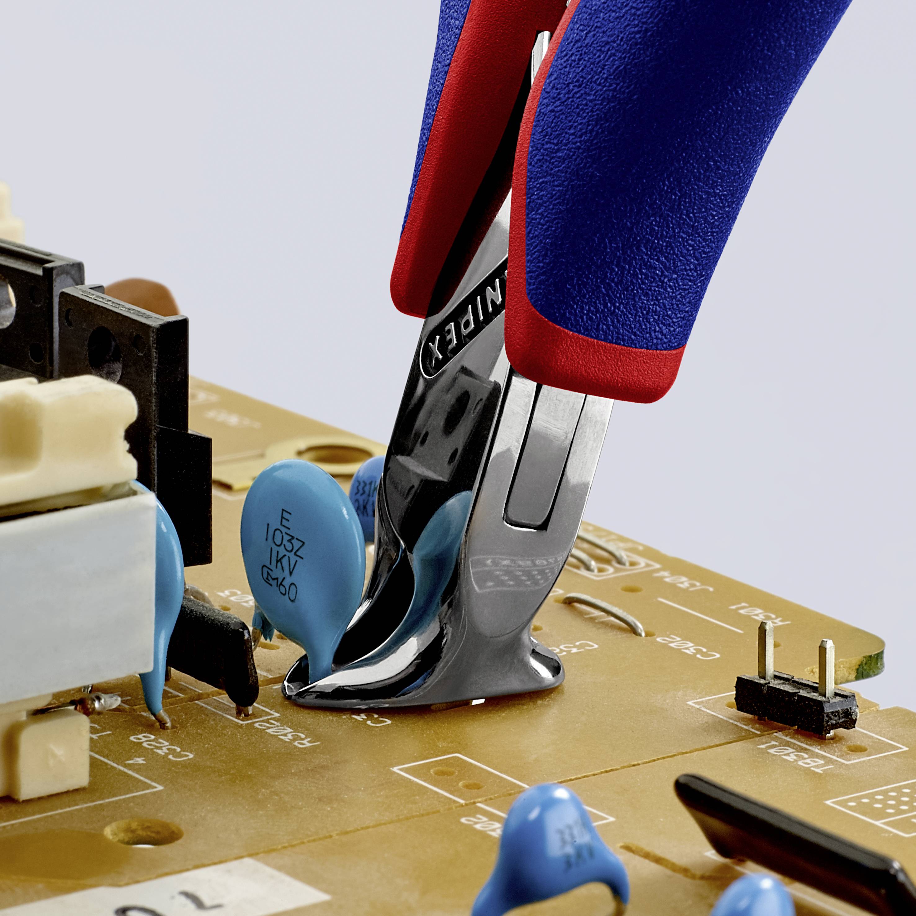 Hand hält rote und blaue Zange, schneidet Draht auf einer Leiterplatte mit verschiedenen elektronischen Bauteilen.
