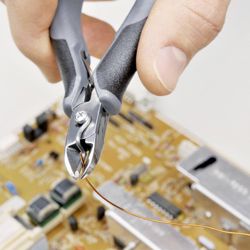 Eine Hand hält einen Drahtschneider, der einen dünnen Draht über einer elektronischen Platine schneidet, die mit verschiedenen Komponenten bestückt ist.