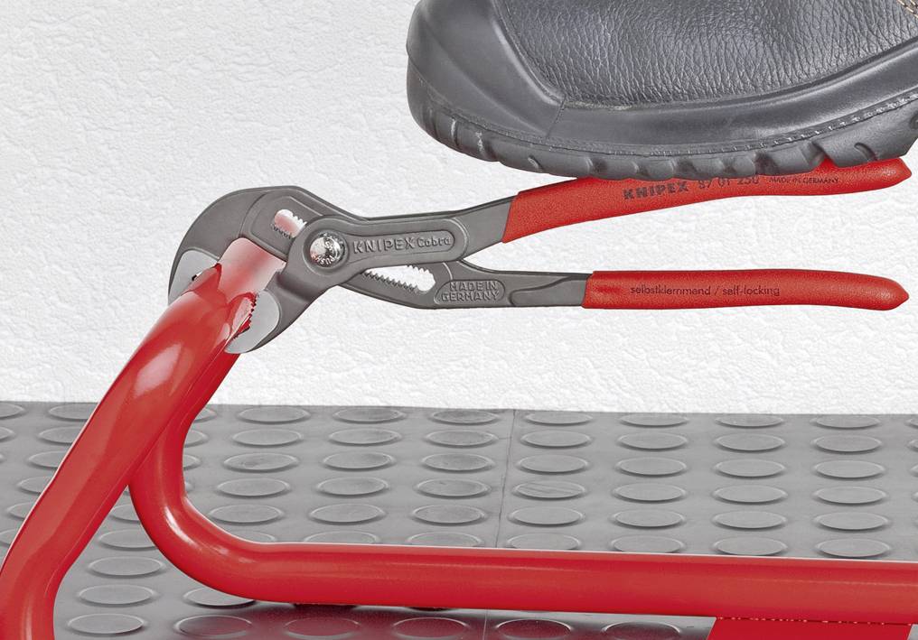 Ein Schuh steht auf einer roten Zange, die ein rohrförmiges Metallteil umfasst, auf einer gepunkteten Bodenplatte.