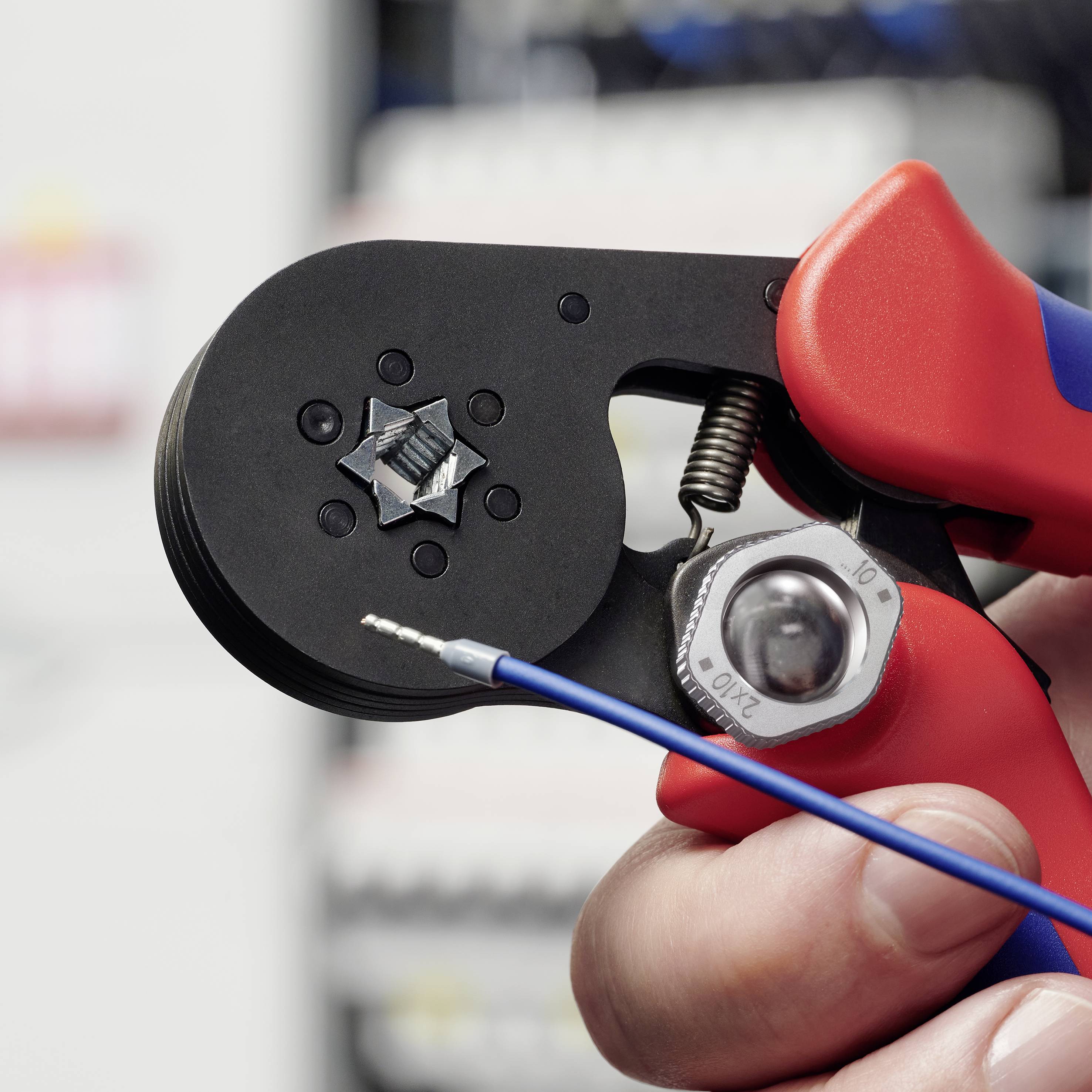 Hand hält eine rote Crimpzange an einem blauen Kabel. Die Zange ist an einem sechseckigen Anschluss positioniert.
