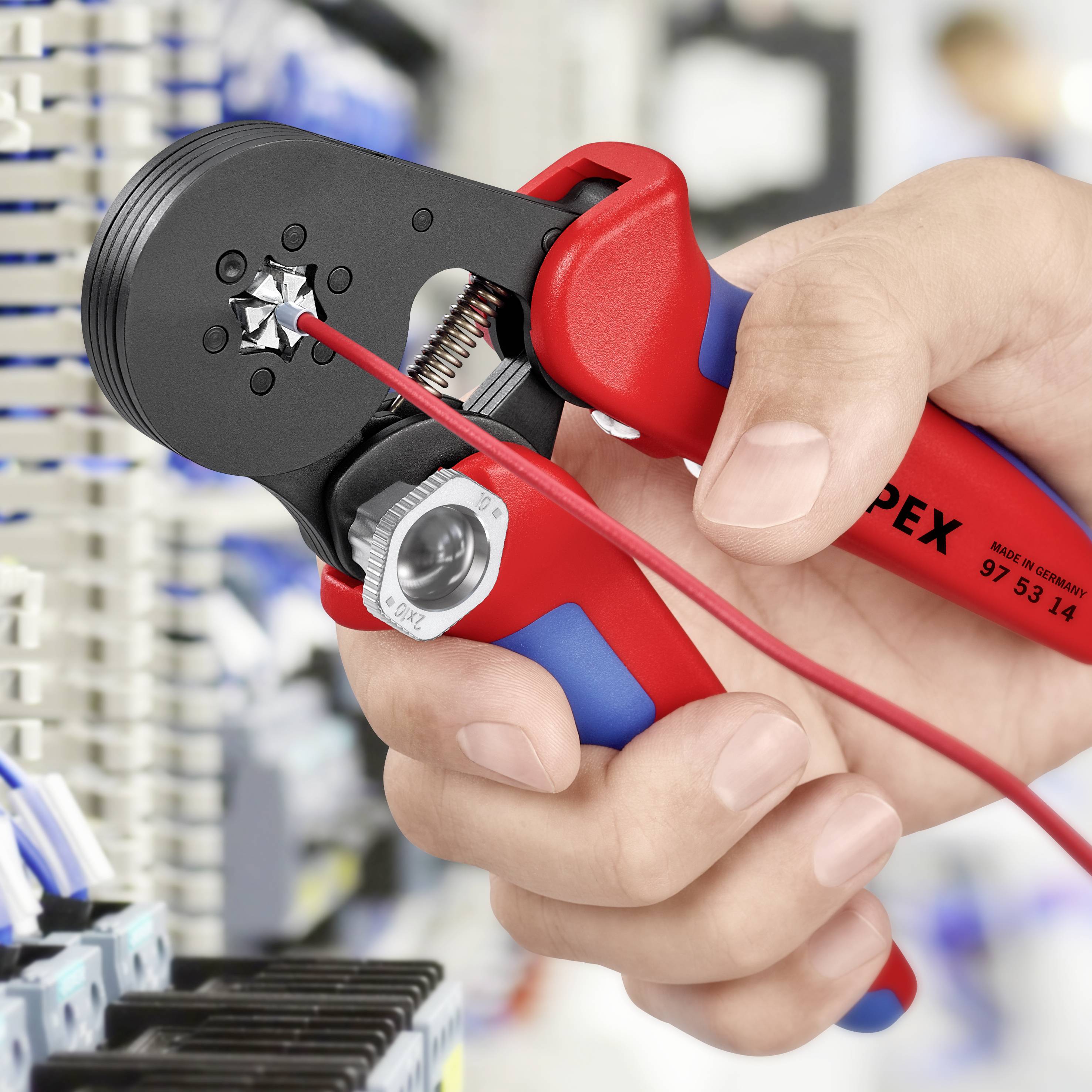 Eine Hand hält eine rote Crimpzange, die ein Kabel bearbeitet. Im Hintergrund ist ein Schaltschrank mit Kabeln zu sehen.