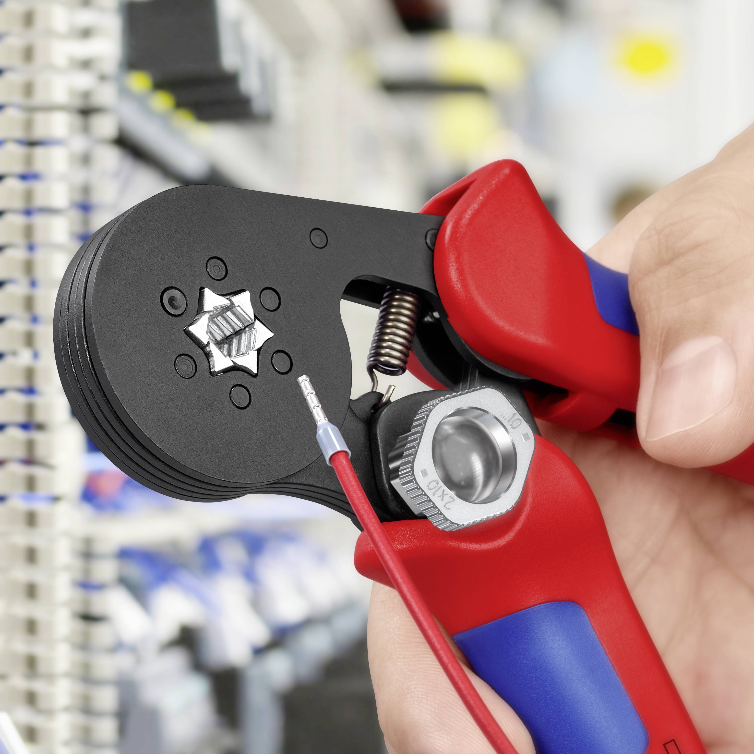 Eine Hand hält eine rote Crimpzange und klemmt einen Metallstecker mit einem Kabel in einem technischen Labor oder Werkstattumfeld.