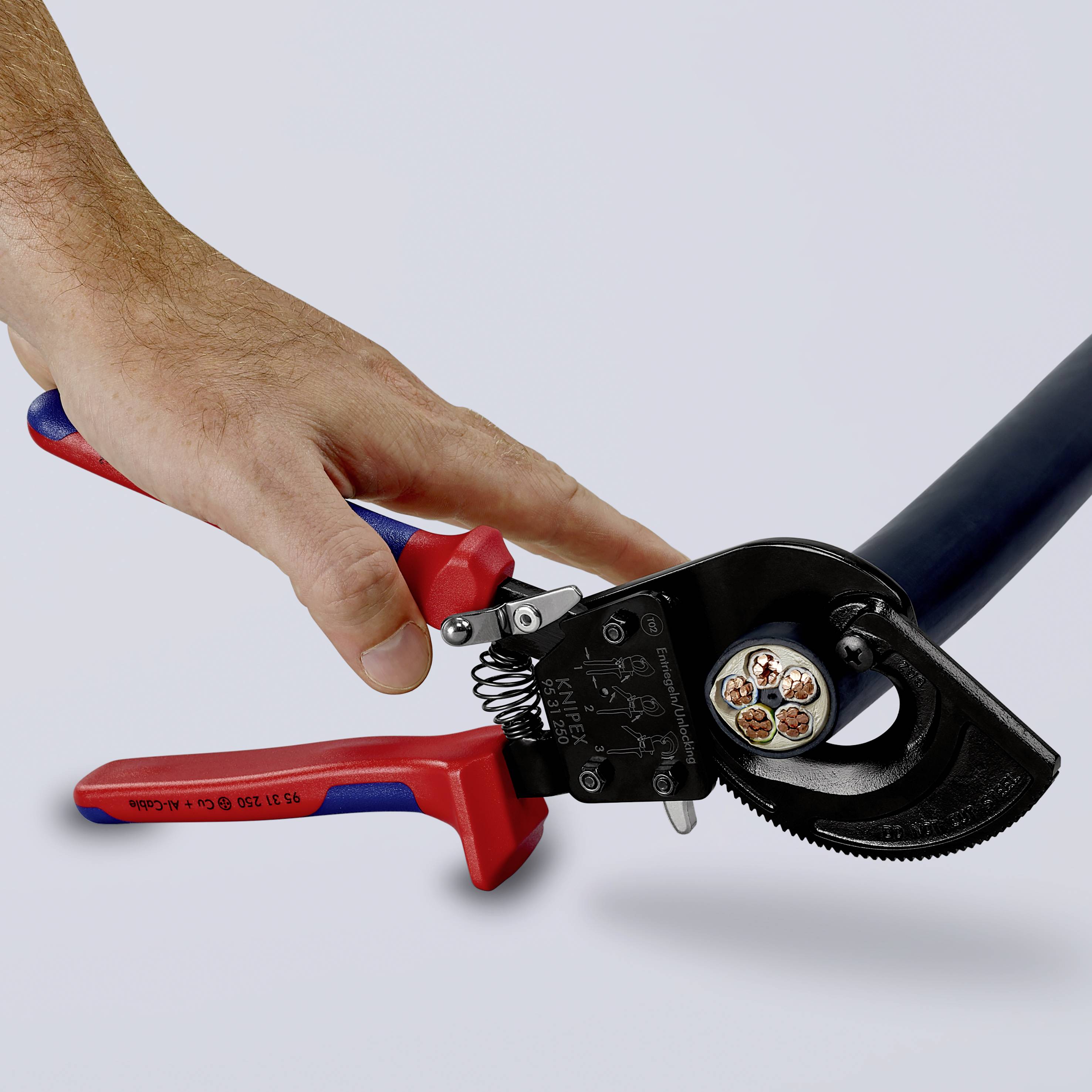 Hand hält einen Kabelschneider, der ein dickes, mehradriges Kabel durchschneidet. Das Werkzeug hat rote und blaue Griffe.