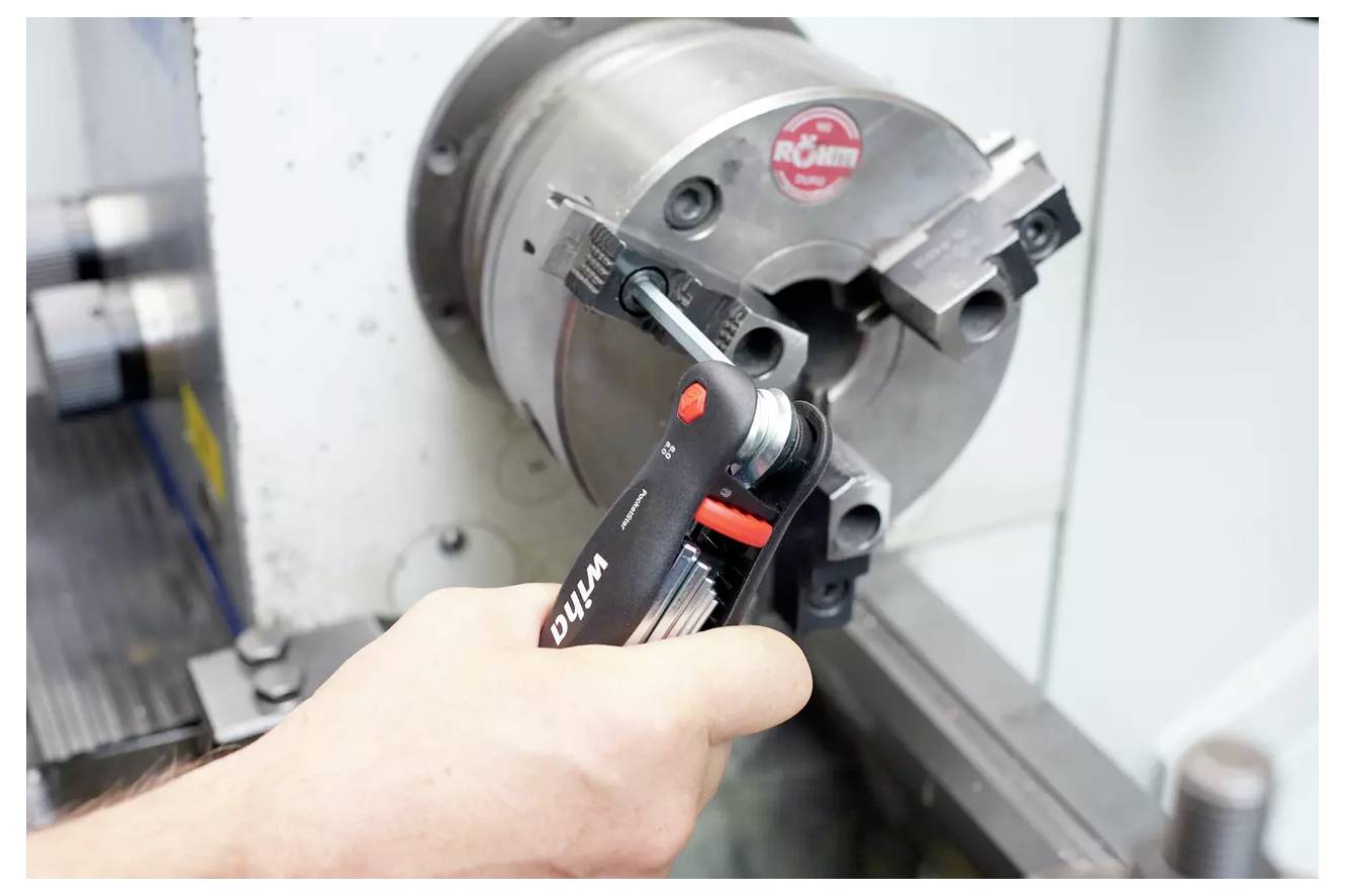 Eine Hand benutzt einen Innensechskantschlüssel, um ein Metallbauteil an einer Drehmaschine in einer Werkstattumgebung einzustellen.