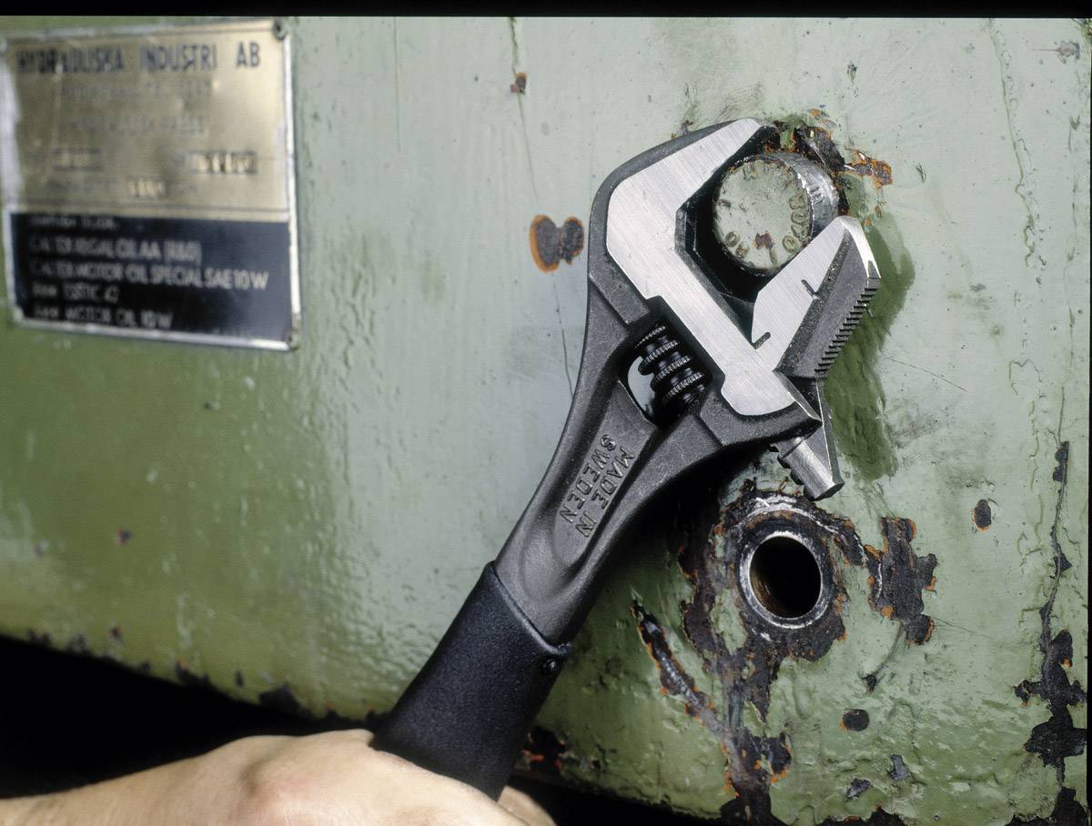 Hand hält verstellbaren Schraubenschlüssel an rostigem Rohr einer grünen Metallfläche.