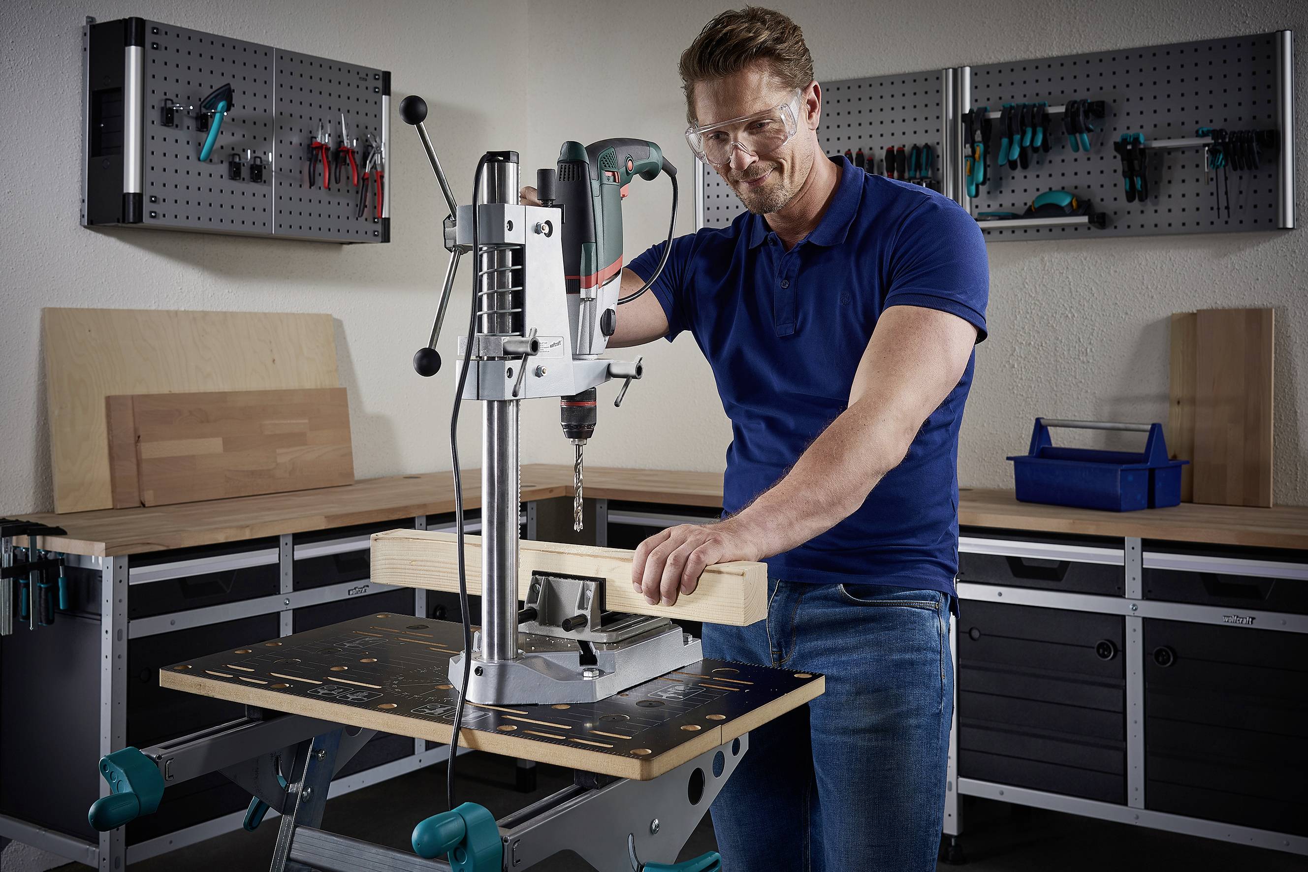Ein Mann in blauer Kleidung nutzt eine Standbohrmaschine, um ein Holzstück in einer Werkstatt zu bearbeiten. Werkzeuge sind im Hintergrund sichtbar.