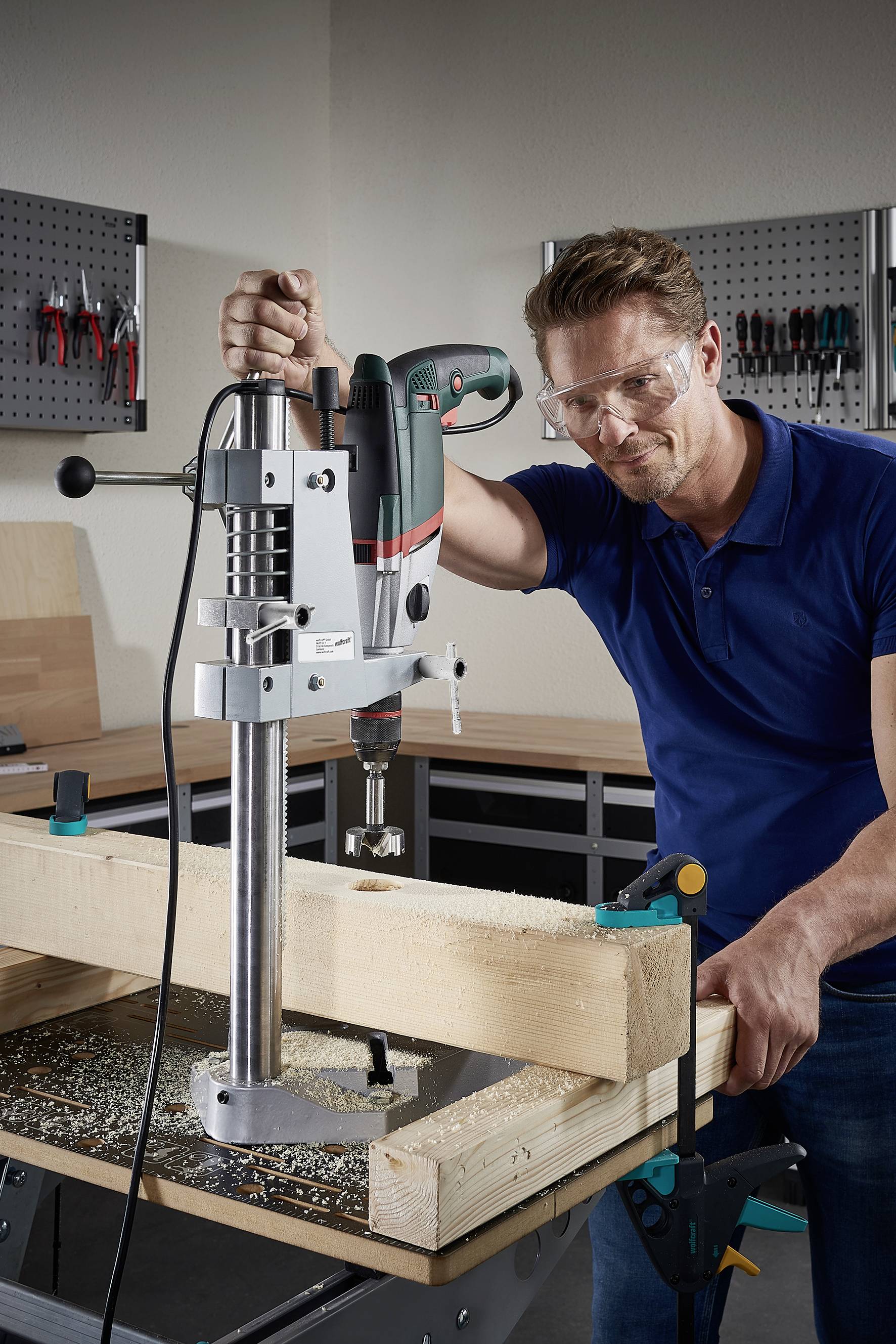 Ein Mann benutzt eine Standbohrmaschine, um in einen großen Holzblock zu bohren. Er trägt Schutzbrille und steht in einer Werkstatt.