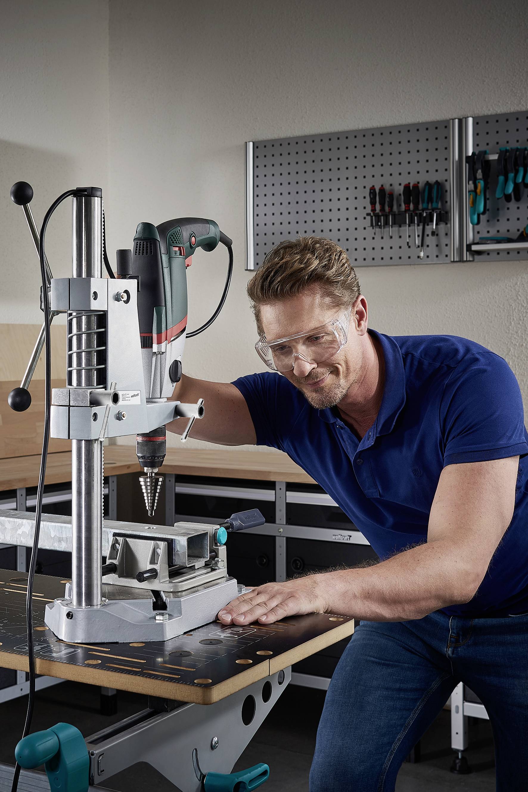 Ein Mann benutzt eine Tischbohrmaschine in einer Werkstatt. Er trägt eine Schutzbrille. Werkzeuge hängen an der Wand im Hintergrund.