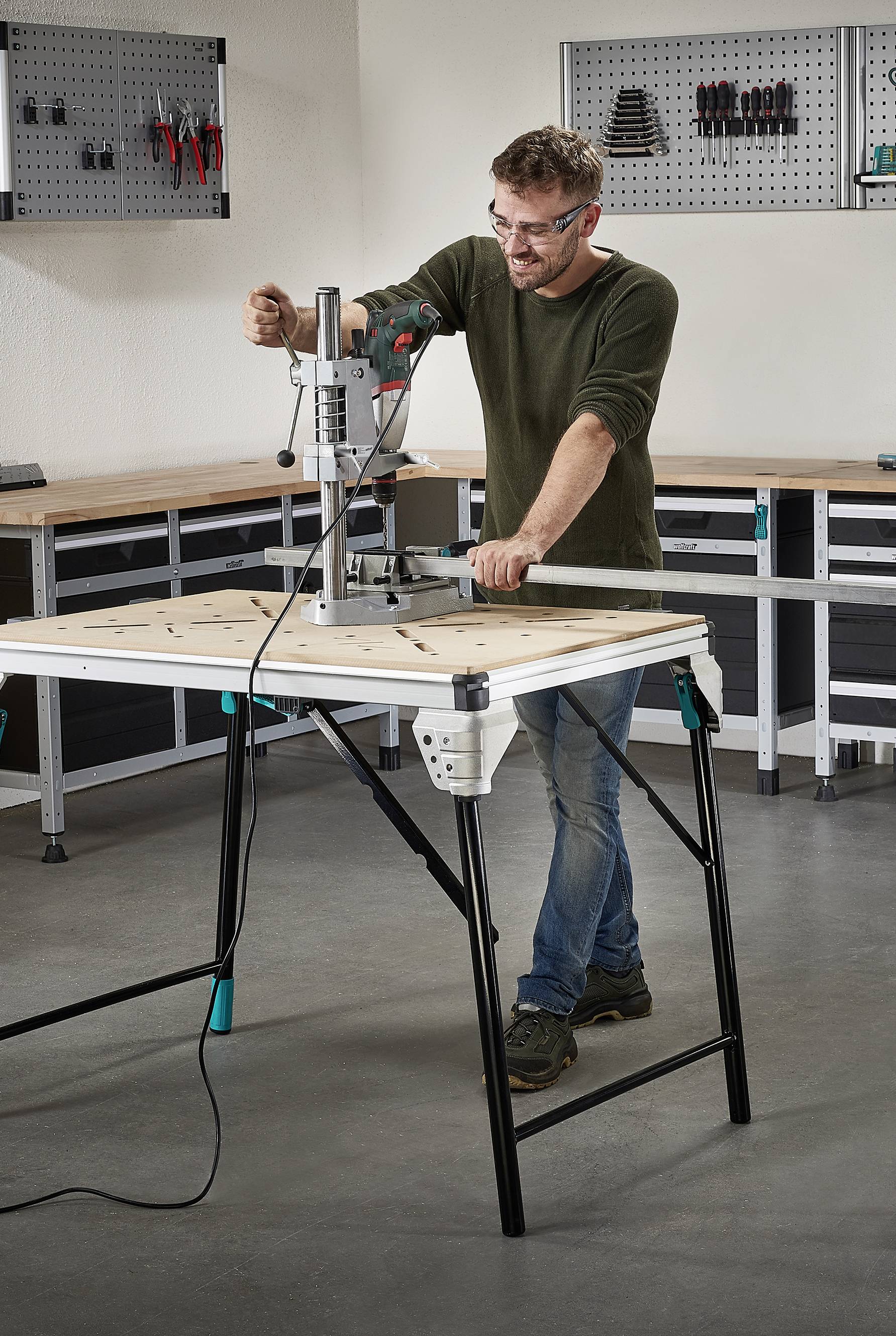 Person bei der Arbeit mit einer Bohrmaschine auf einem Montagetisch in einer Werkstatt. Werkzeuge sind an der Wand im Hintergrund sichtbar.