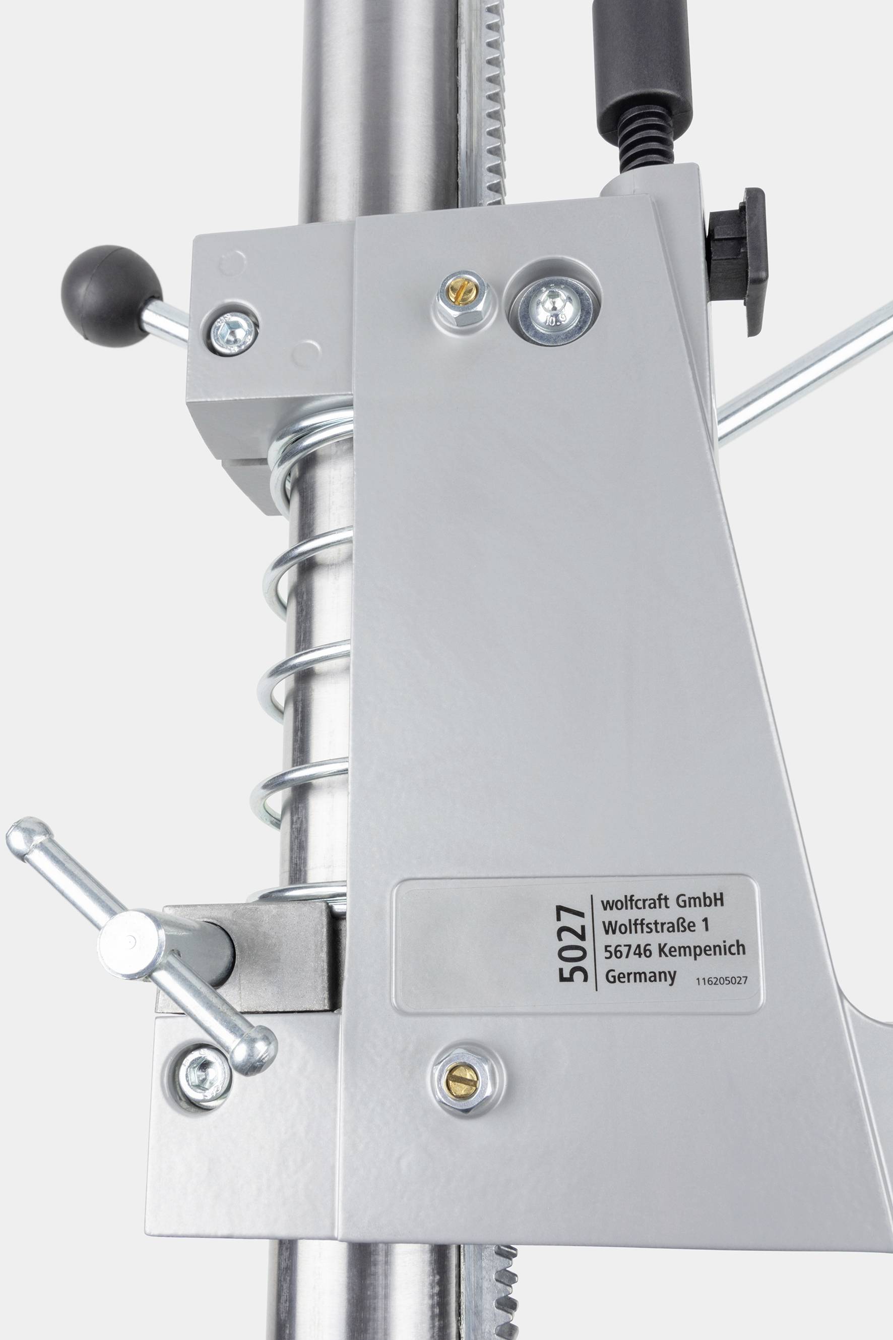 Maschinenkomponente mit Metallhebel und Feder, etiketiert mit '5027' und Adresse von 'wolfcraft GmbH, Kempenich, Germany'.