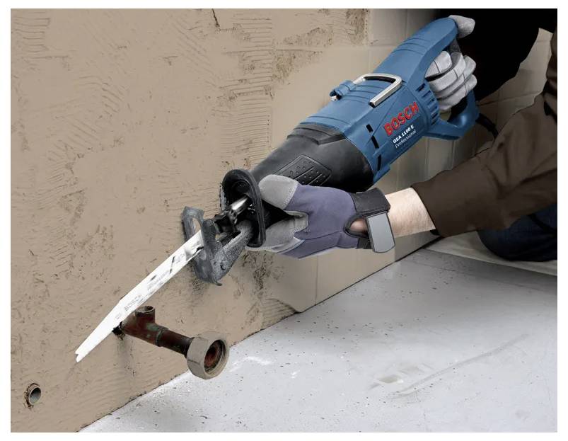 Eine Person benutzt eine Bosch-Säbelsäge, um durch eine braune Wand zu schneiden, wobei sie auf ein Metallrohr abzielt. Die Person trägt Handschuhe aus Sicherheitsgründen.