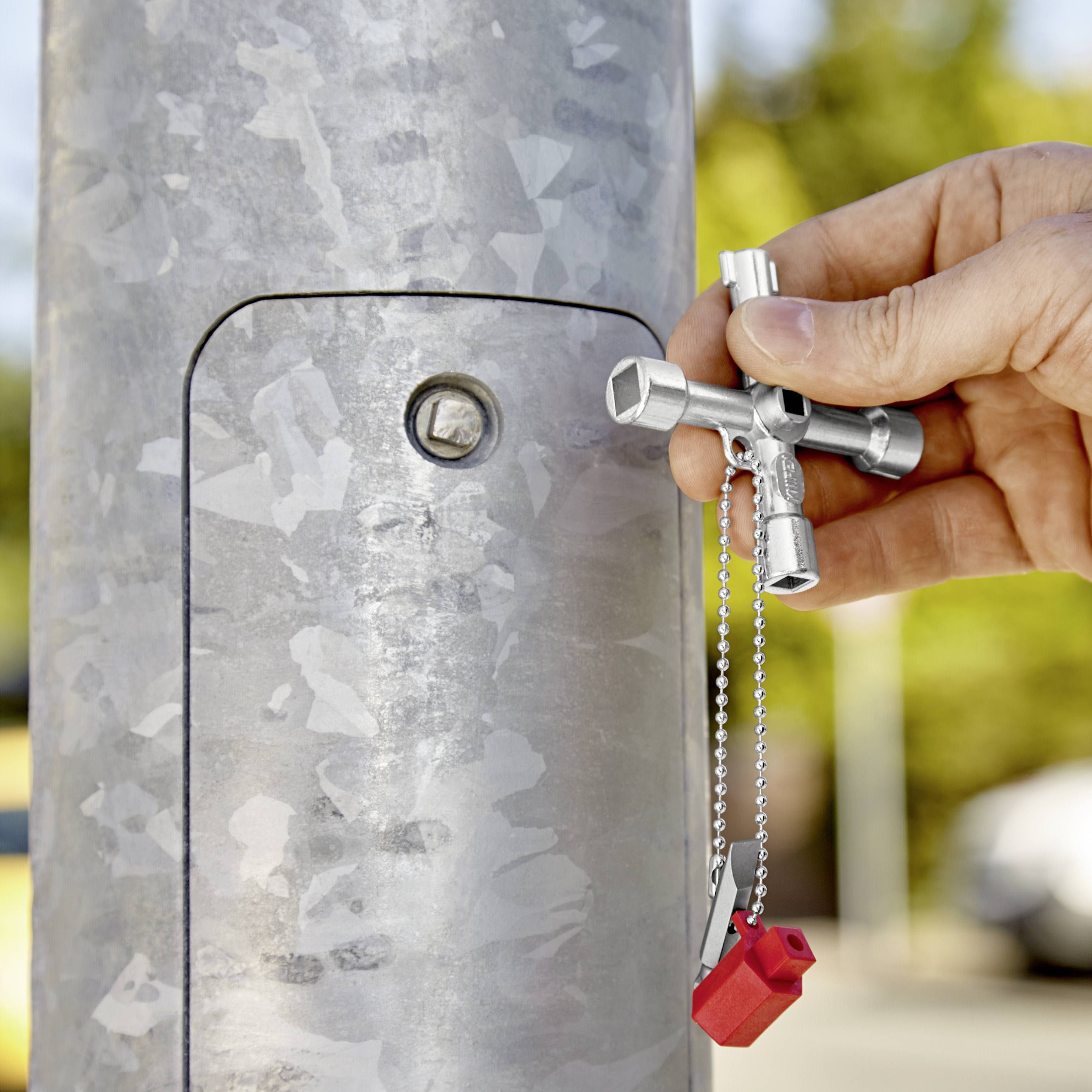 Eine Hand hält einen silbernen Vierkantschlüssel, um einen Verschluss an einem Metallpfosten zu öffnen. Daneben hängt ein rotes Spielzeughaus.
