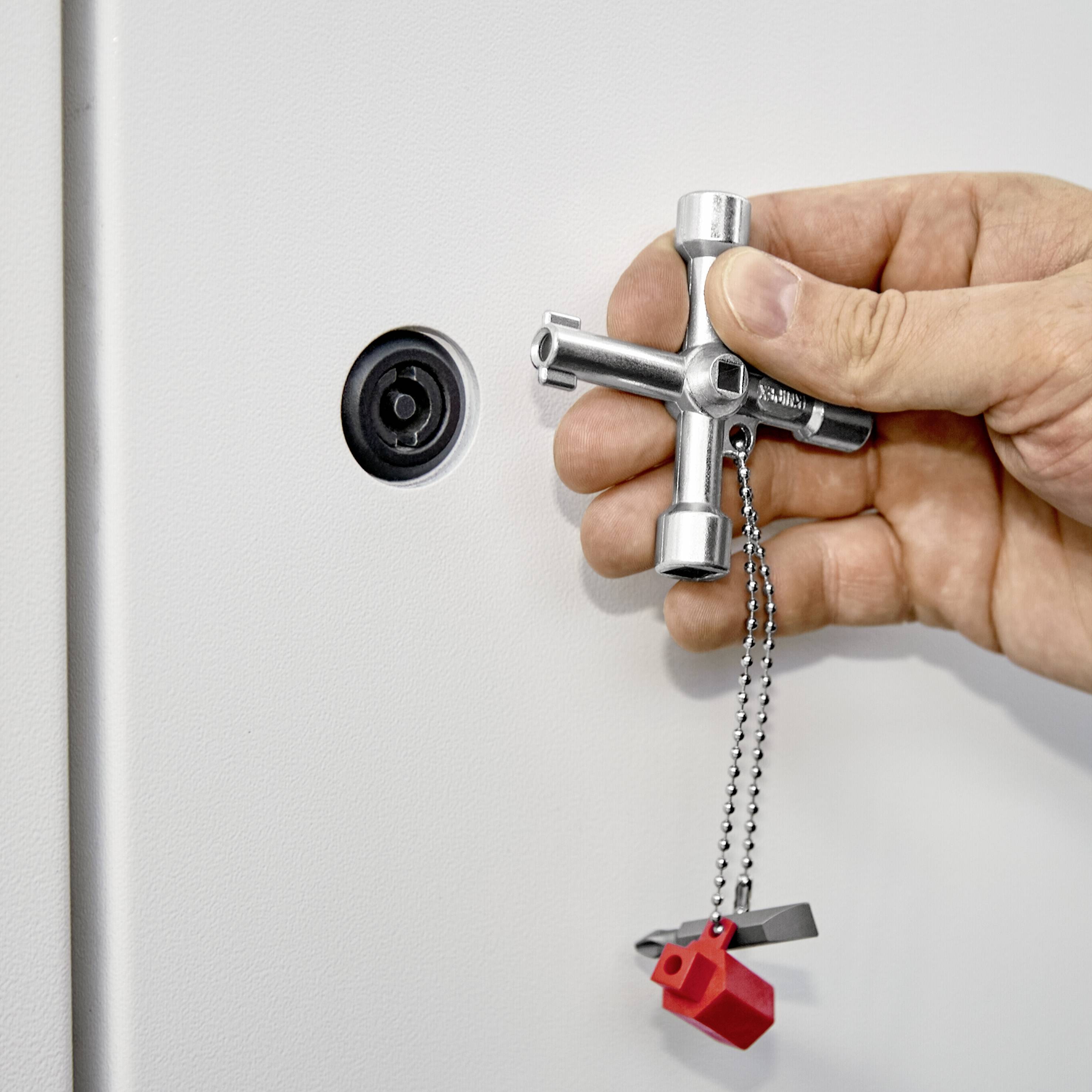 Eine Hand hält einen Kreuzschlüssel, um ein Schließfach zu öffnen. Der Schlüssel hängt an einer Kette mit einem roten Plastikanhänger.