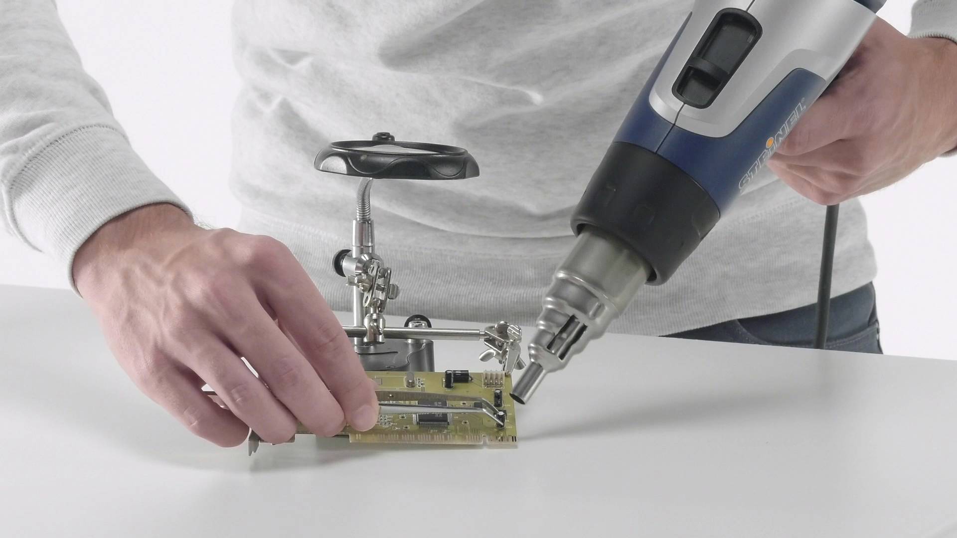 Eine Person lötet eine Leiterplatte mit einem Heißluftgerät. Sie hält die Platine auf einem Halter, während das Gerät in der anderen Hand ist.