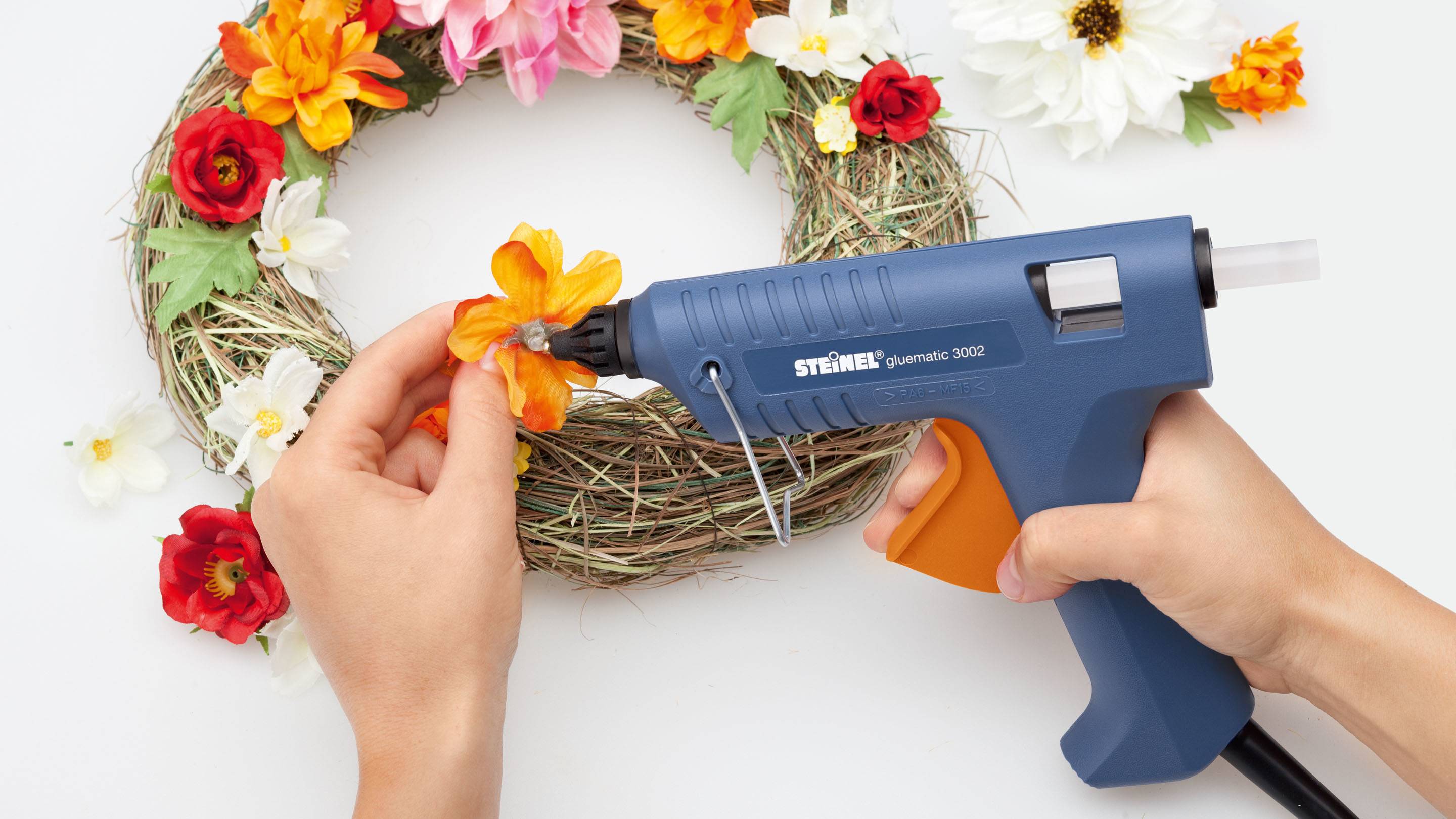 Eine Person befestigt orangefarbene Blume mit Klebepistole an Kranz aus Zweigen und Blumen. Feierliche Dekoration wird erstellt.