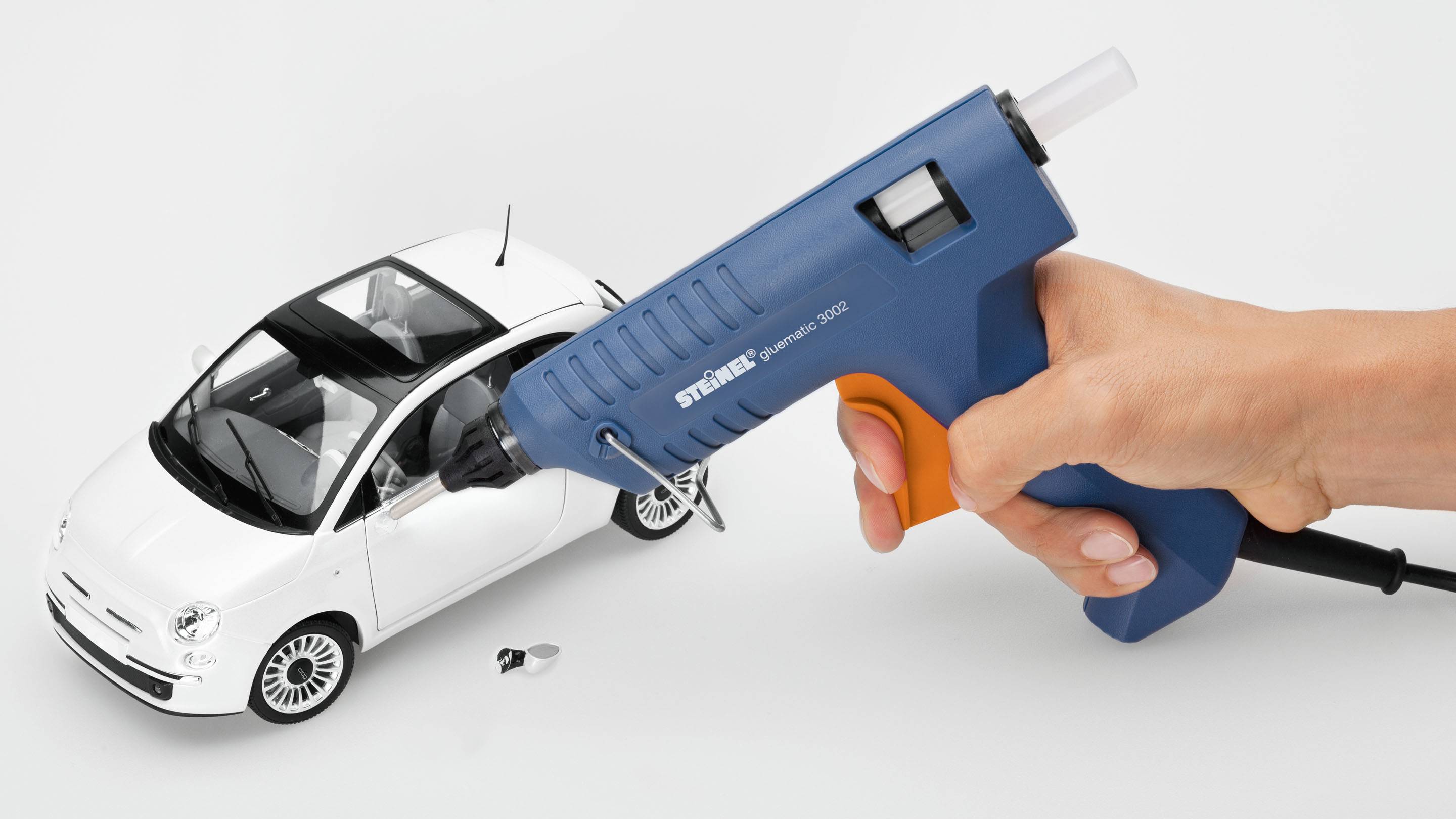 Jemand hält eine blaue Heißklebepistole nahe eines weißen Modellautos, als ob das Auto repariert wird.