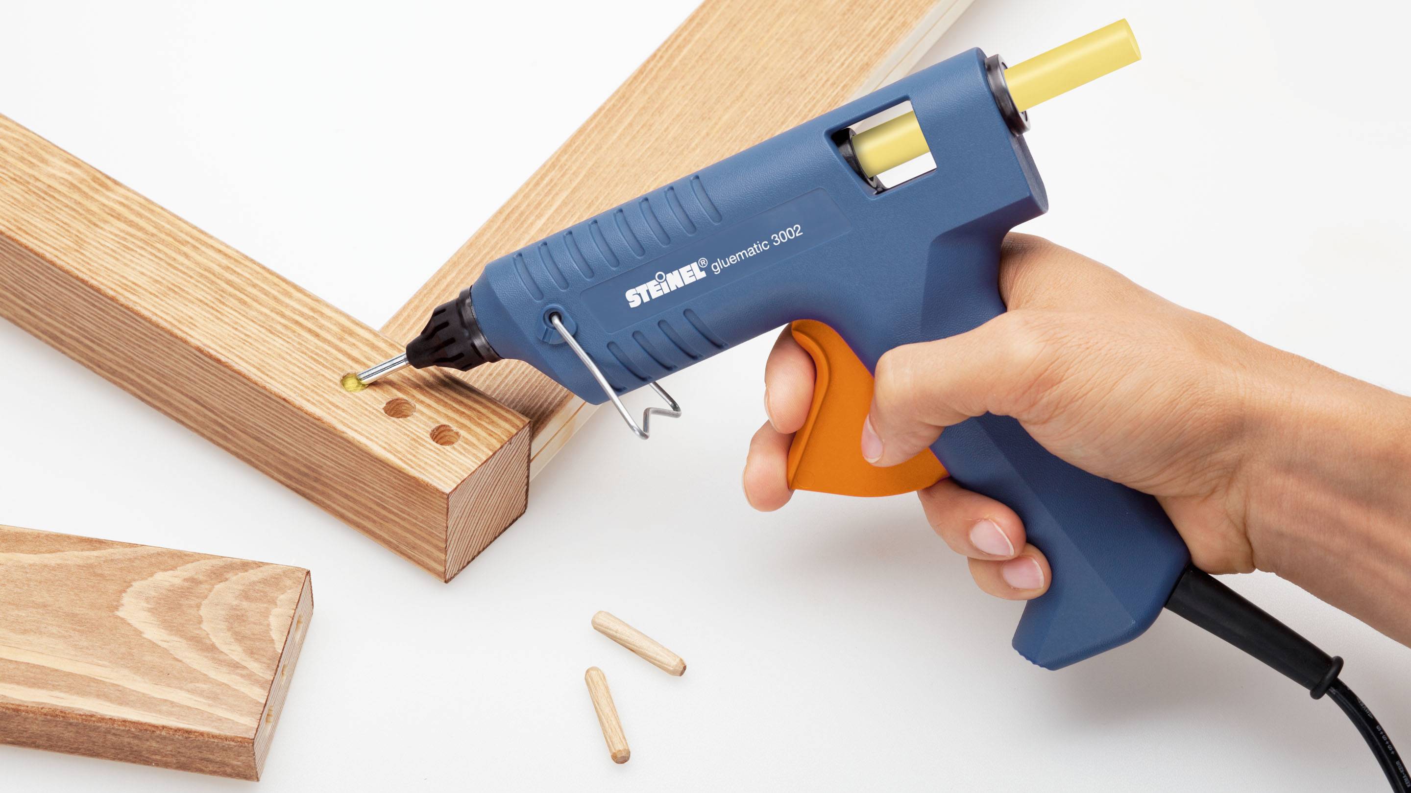 Eine Hand hält eine blaue Heißklebepistole mit gelbem Klebestift und verklebt zwei Holzstücke miteinander.