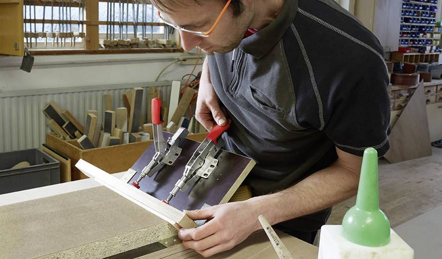 Ein Mann arbeitet in einer Werkstatt und befestigt Holzstücke mit einer Zwinge. Im Hintergrund sind Regale mit weiteren Materialien sichtbar.