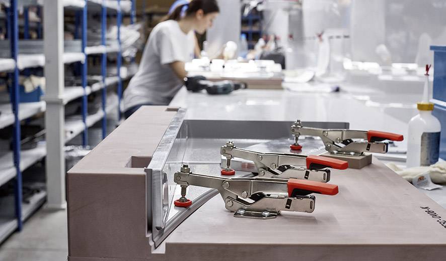 Eine Werkstatt mit Fokus auf drei Metallklemmen, die ein Materialstück halten; im Hintergrund verschwommen eine Person bei der Arbeit.