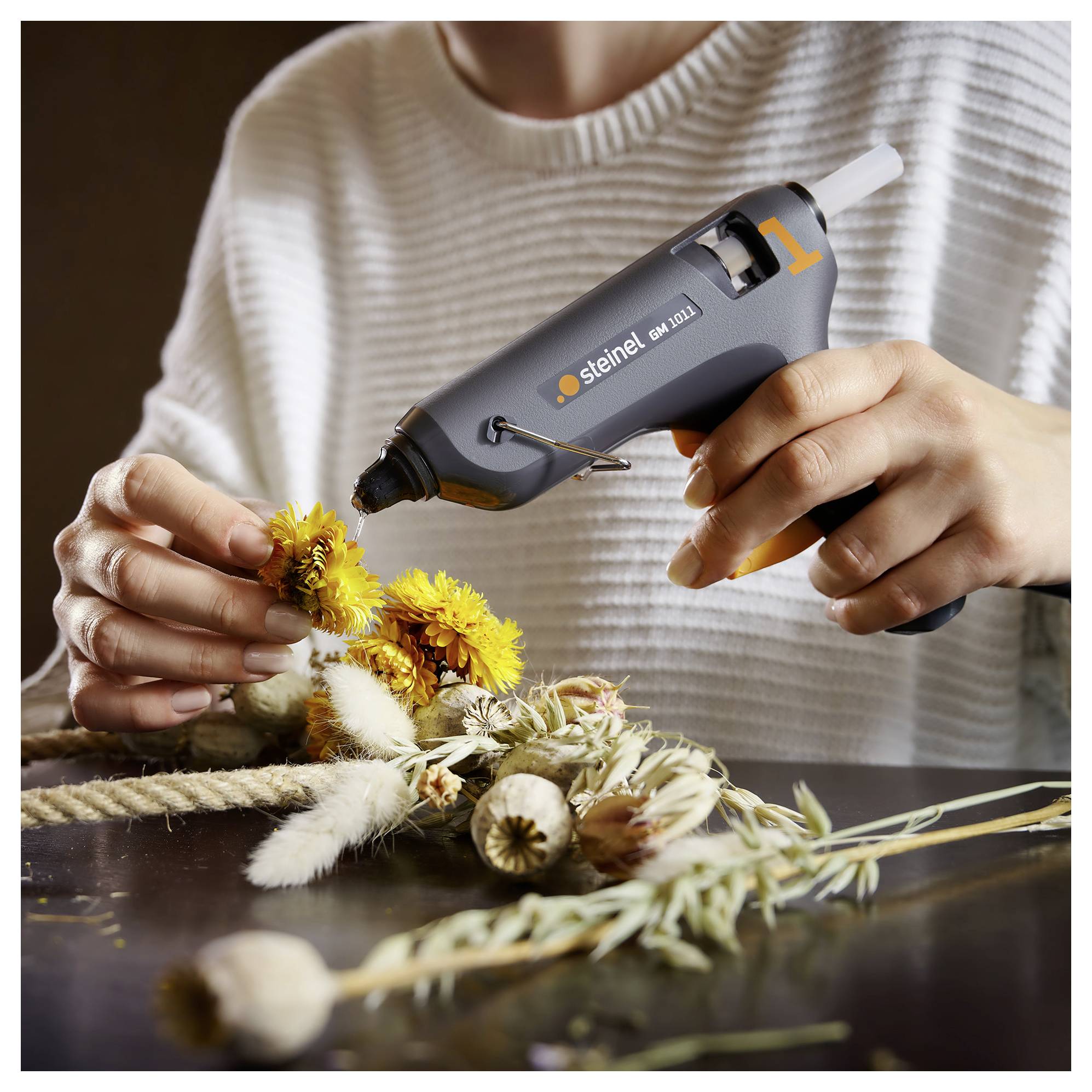 Hände verwenden eine Heißklebepistole, um getrocknete Blumen und Gräser auf eine Bastelgestaltung auf einem Tisch zu befestigen.