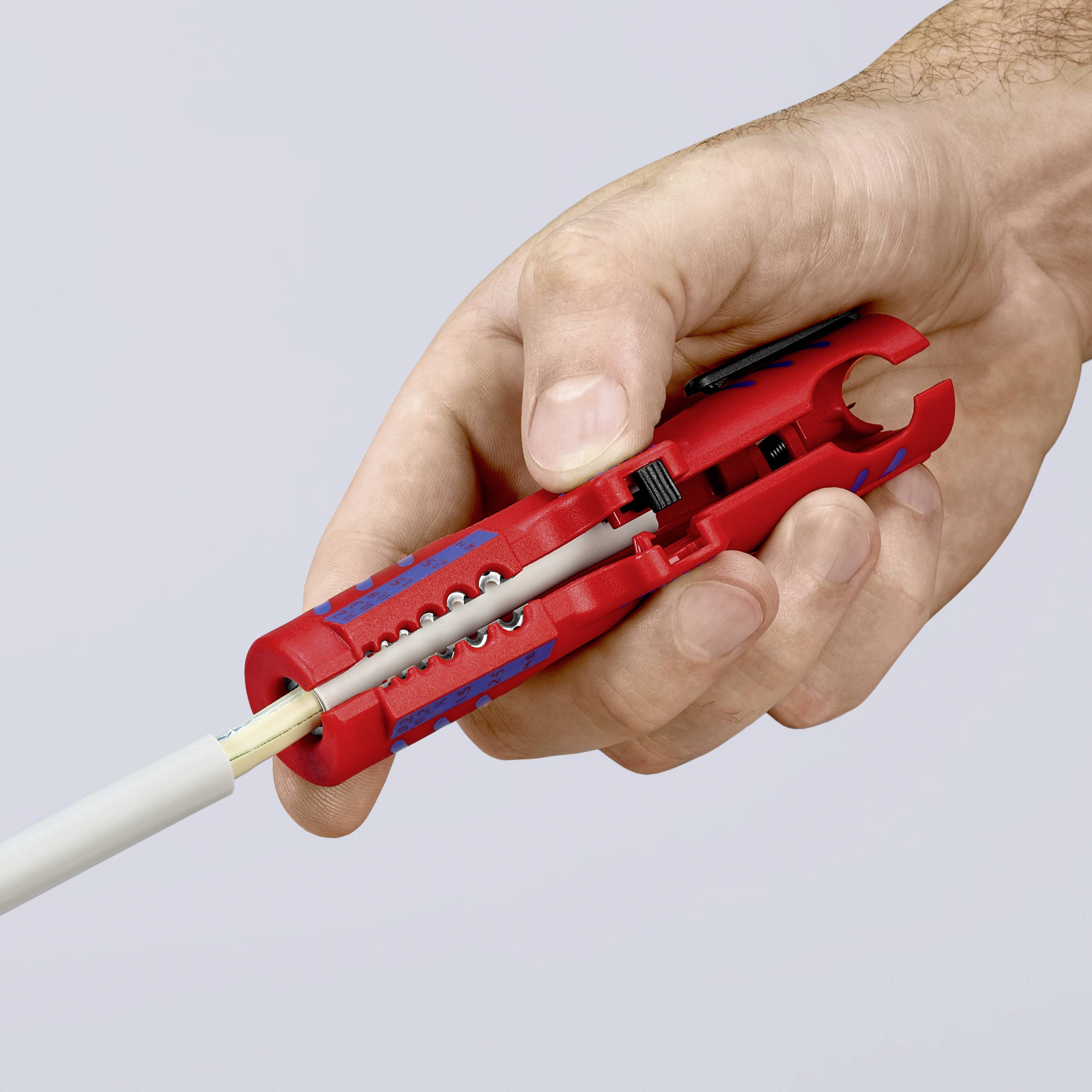 Eine Hand hält ein rotes Abisolierwerkzeug, das ein weißes Kabel umschließt. Das Werkzeug entfernt die Isolierung vom Kabel.