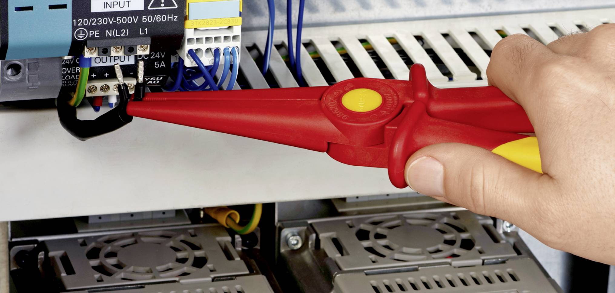 Eine rote Zange hält das schwarze Kabel an einem elektrischen Gerät in einem Schaltschrank. Sicherheitsüberprüfung wird durchgeführt.