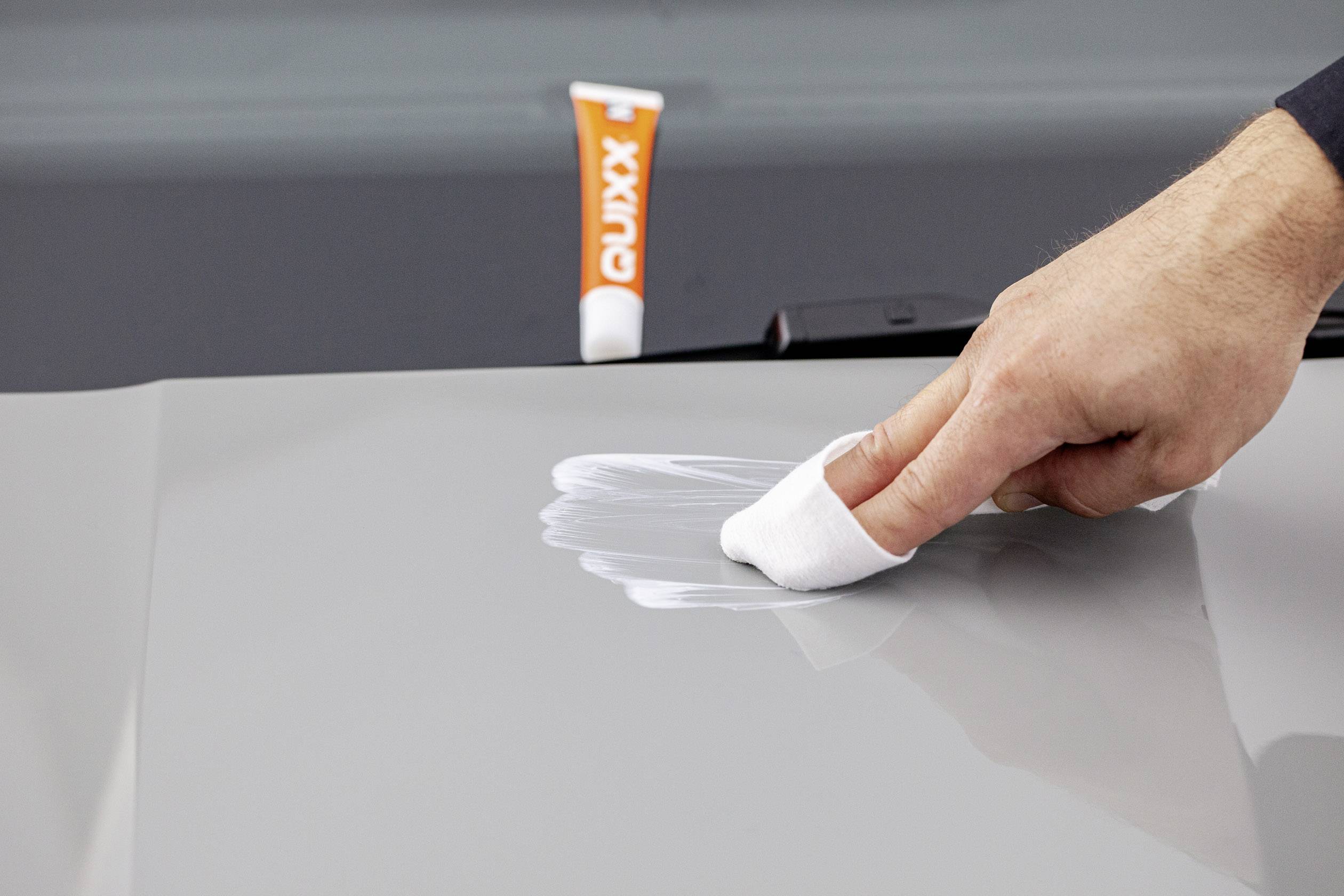 Eine Hand poliert Autolack mit einem weißen Tuch, Tube Autopflegemittel im Hintergrund.
