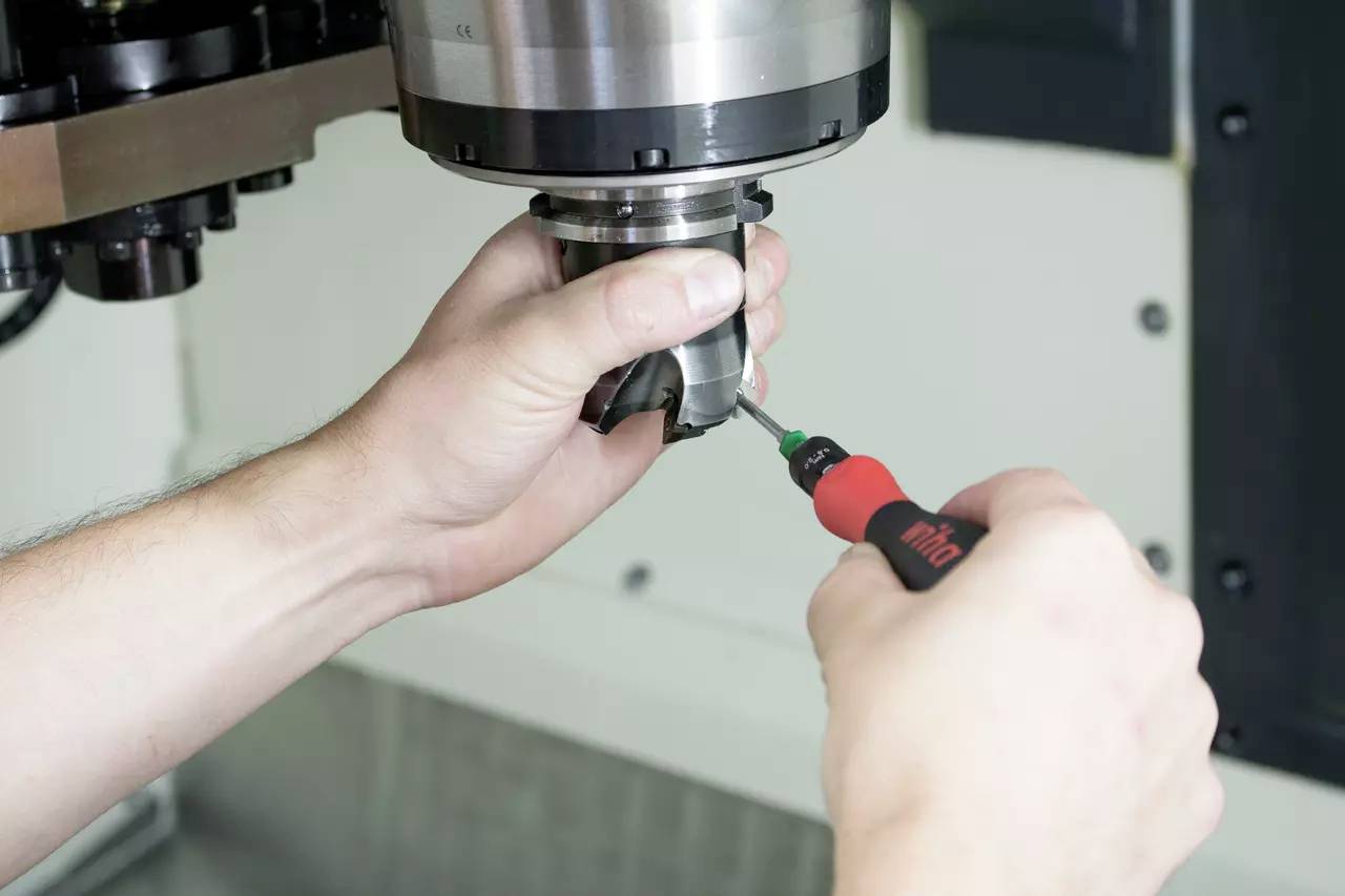 Zwei Hände arbeiten an einer Maschine, eine hält ein Werkzeug, die andere eine Art Schraubenzieher, der auf das Werkzeug gerichtet ist.