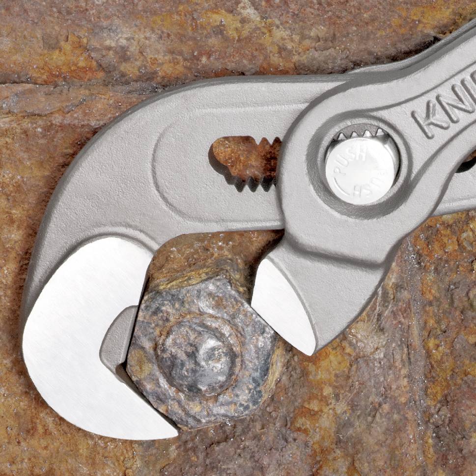 Ein Multifunktionswerkzeug schneidet einen verrosteten Schraubenkopf auf einem roten Steinboden. Werkzeug besteht aus Metall.