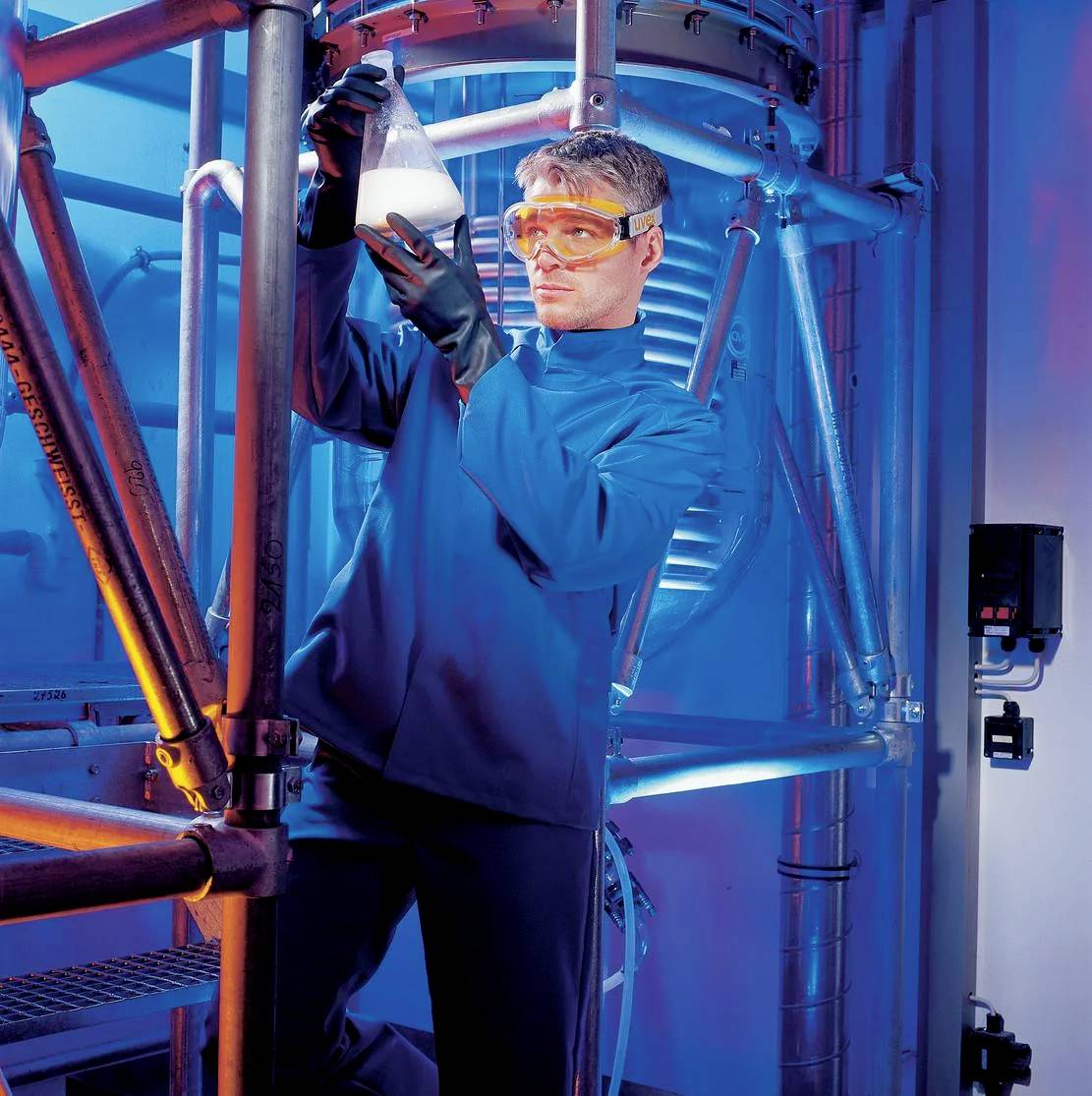 Ein Mann in Schutzkleidung und Schutzbrille hält ein Reagenzglas in einer industriellen Umgebung mit blauer Beleuchtung.