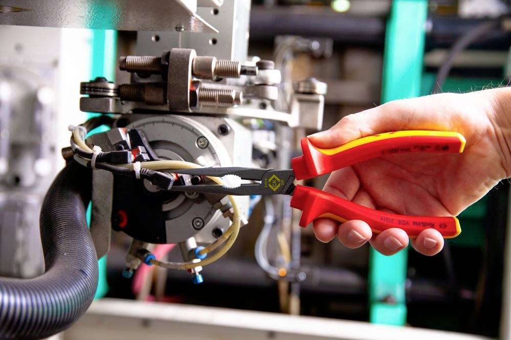 Eine Hand hält eine rote Zange, die an einem technischen Gerät arbeitet. Im Hintergrund sind Maschinen und Kabel sichtbar.