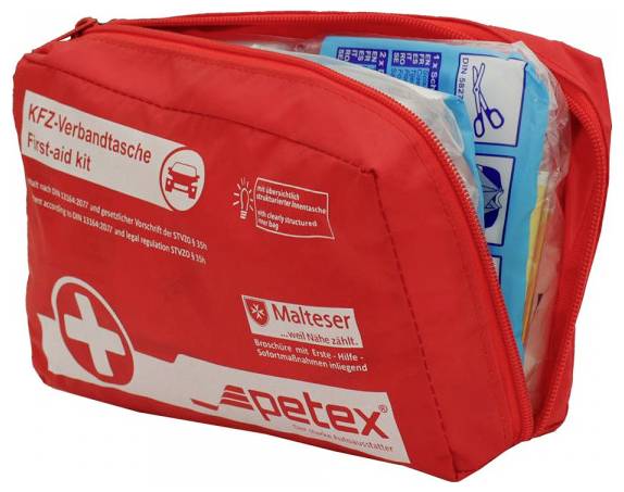 Eine rote Erste-Hilfe-Tasche mit einem Kreuz-Symbol, beschriftet als 'KFZ-Verbandtasche First-aid kit.' Sie ist teilweise geöffnet und zeigt Scheren und Erste-Hilfe-Materialien.