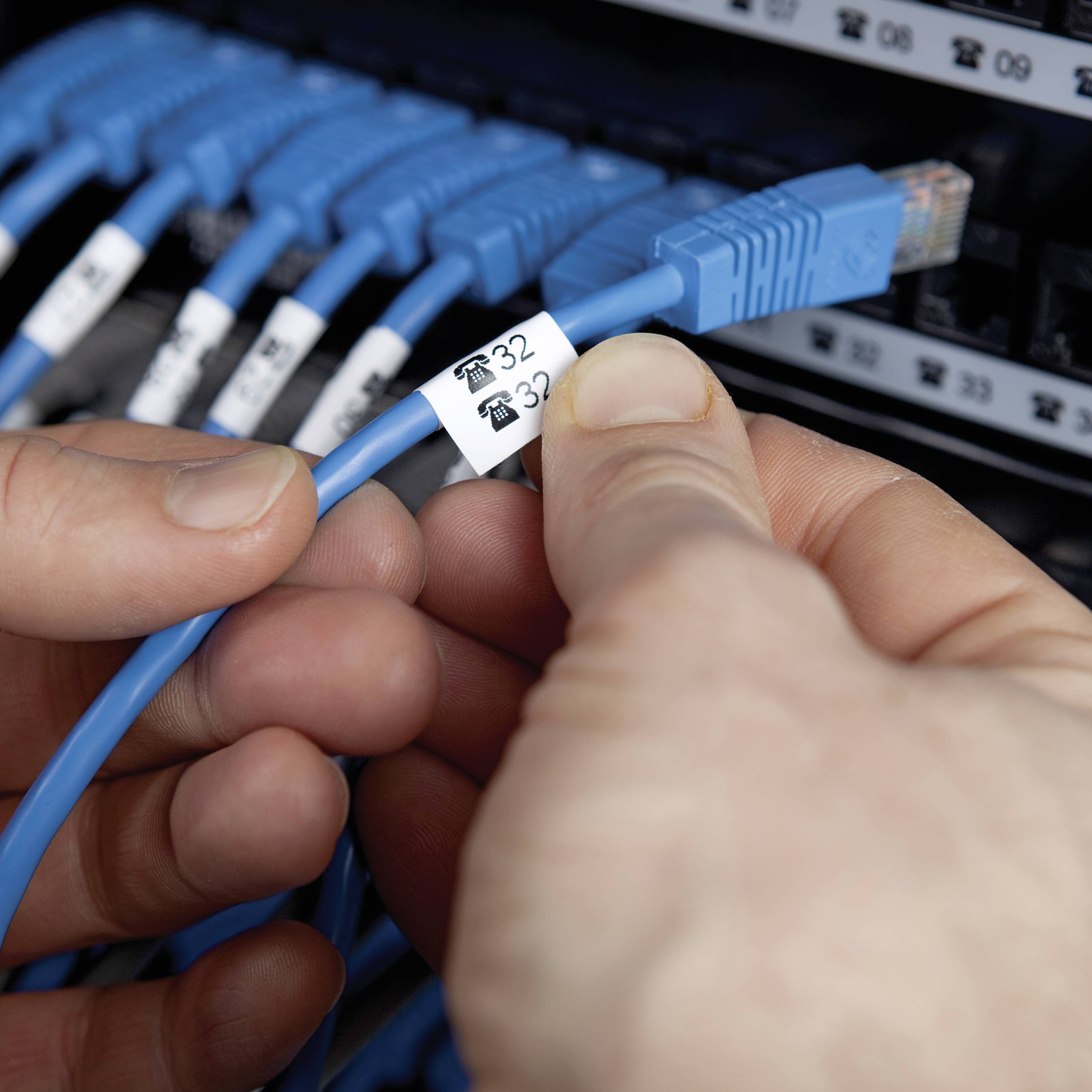 Ein Mensch hält ein blaues Netzwerkkabel mit der Aufschrift '32' vor einem Server-Rack. Mehrere blaue Kabel sind angeschlossen.