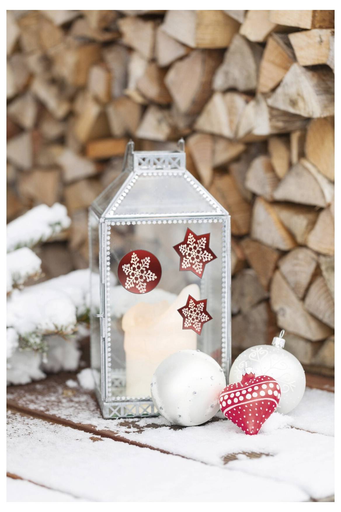 Eine leuchtende silberne Laterne mit roten Schneeflockenornamenten und weißen sowie roten Weihnachtskugeln steht auf verschneitem Holz vor einer aufgestapelten Holzwand.