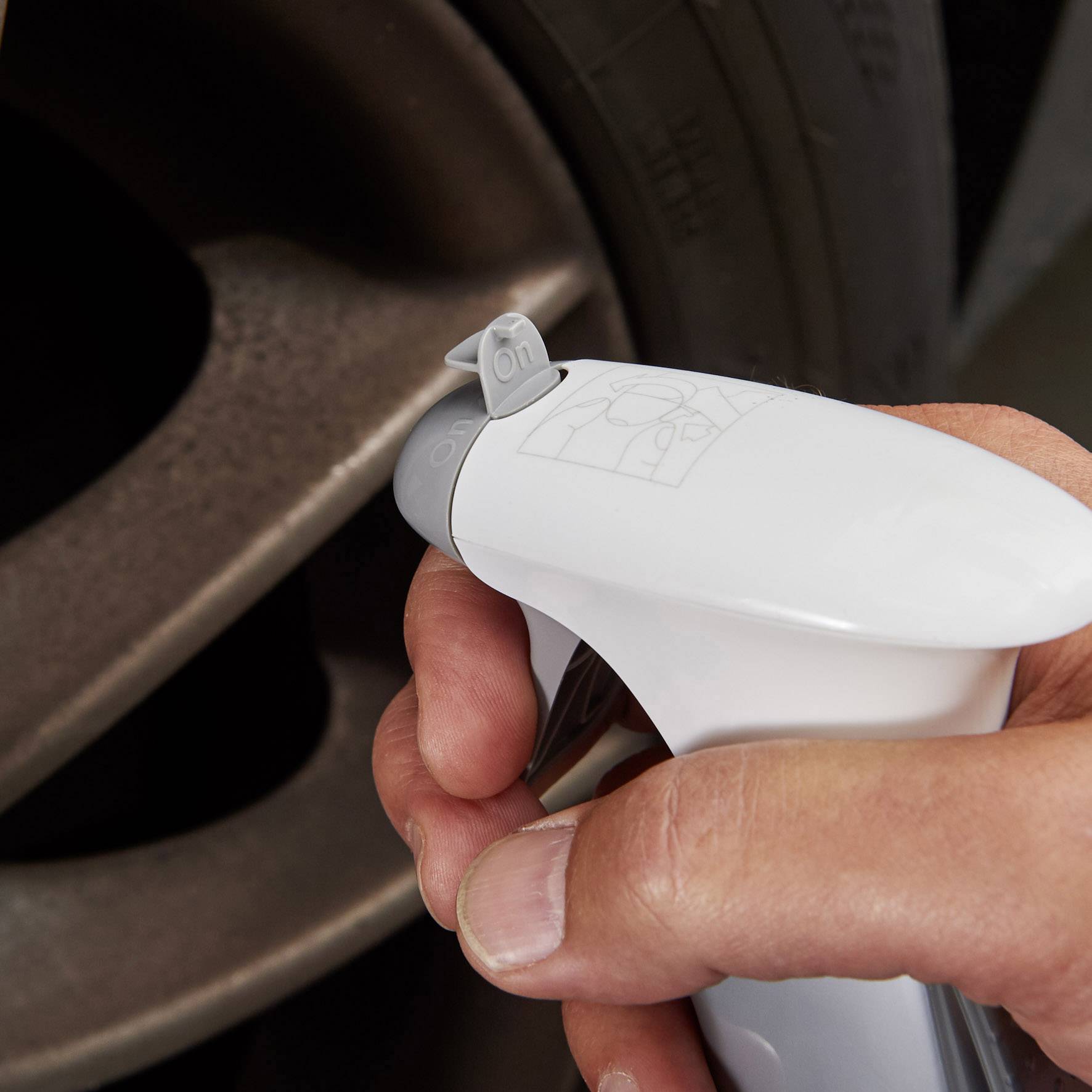 Hand hält eine weiße Sprühflasche, die auf ein Autoreifenventil gerichtet ist, zur Reinigung oder Wartung, mit sichtbarem Ventildeckel.