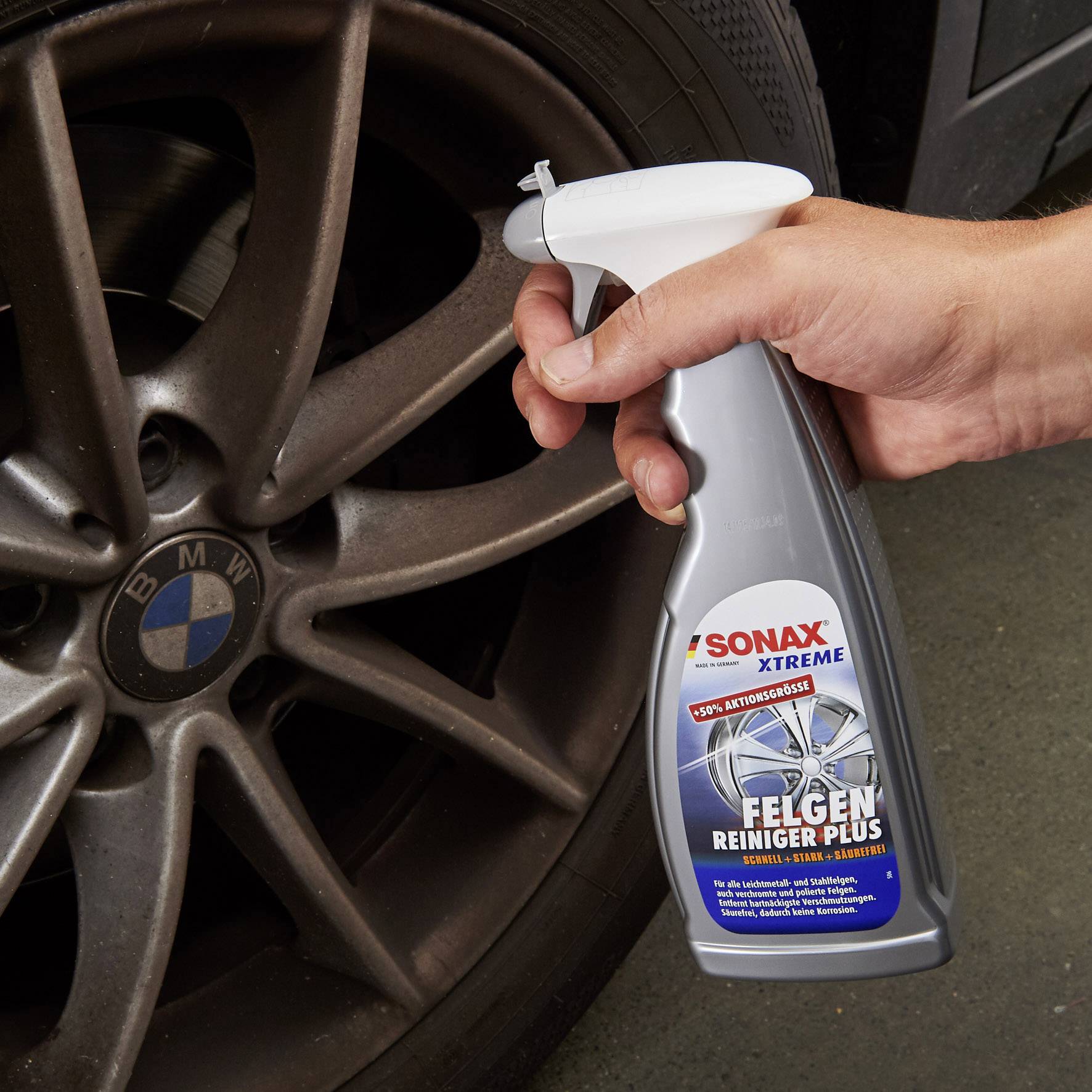 Eine Hand hält einen 'SONAX XTREME' Felgenreiniger vor einem Autoreifen mit BMW-Logo, bereit zur Reinigung der Felgen.