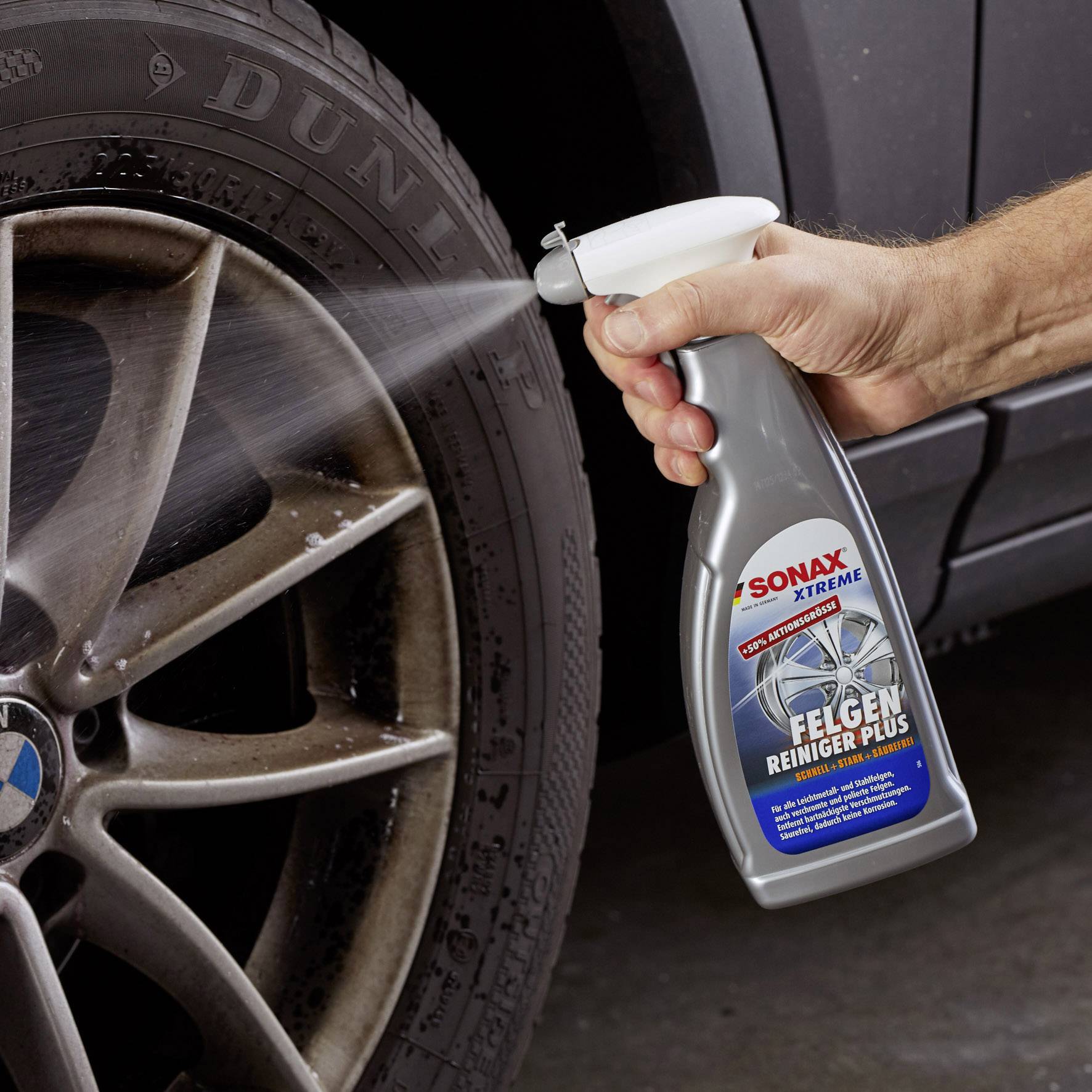 Eine Hand sprüht 'Sonax Xtreme Felgenreiniger Plus' auf ein Autoreifenrad, das sichtbar verschmutzt ist. Im Hintergrund ist ein Autoreifen.