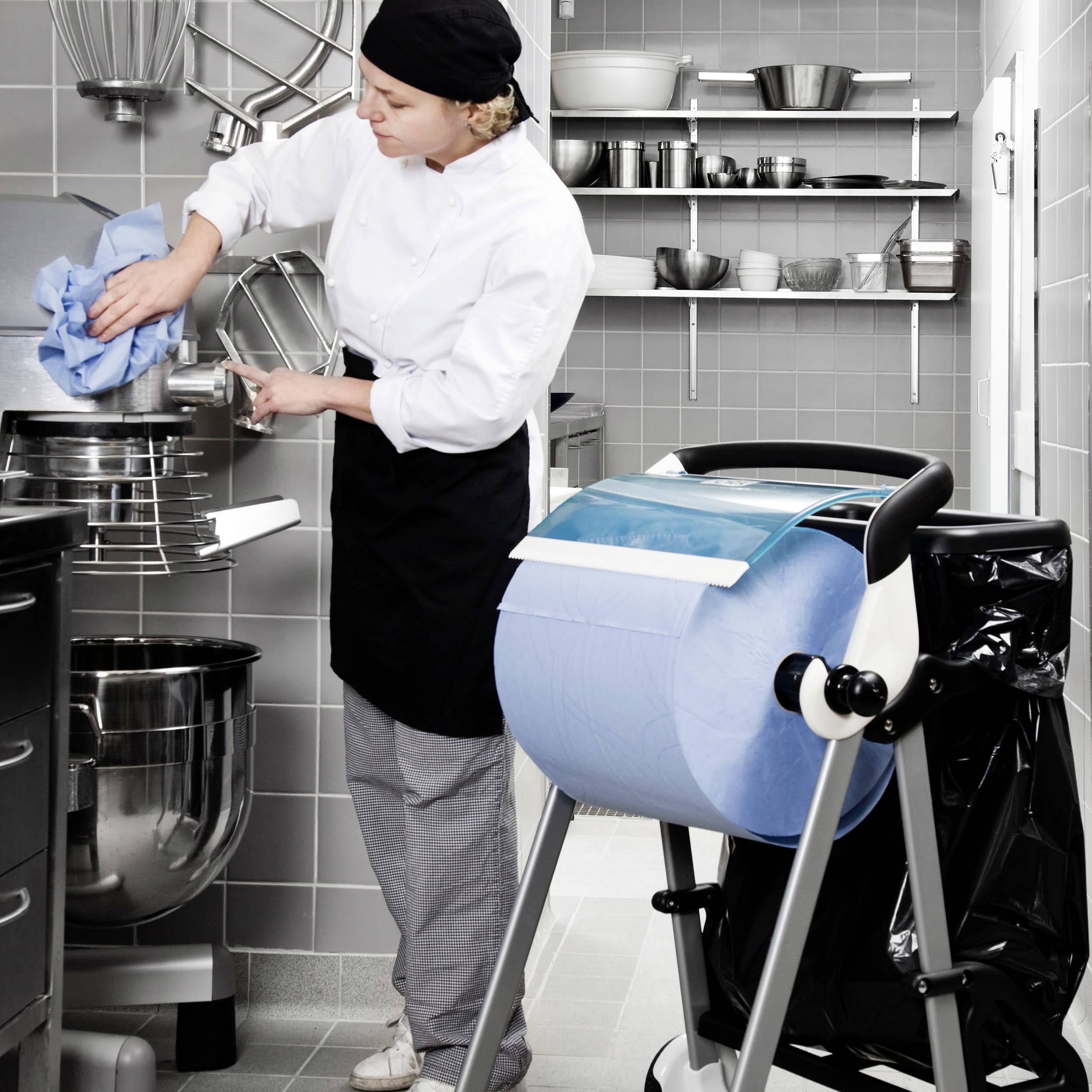 Küchenarbeiterin in weißem Hemd und schwarzer Schürze reinigt einen großen Rührbesen. Im Hintergrund schimmert silbernes Küchengerät.