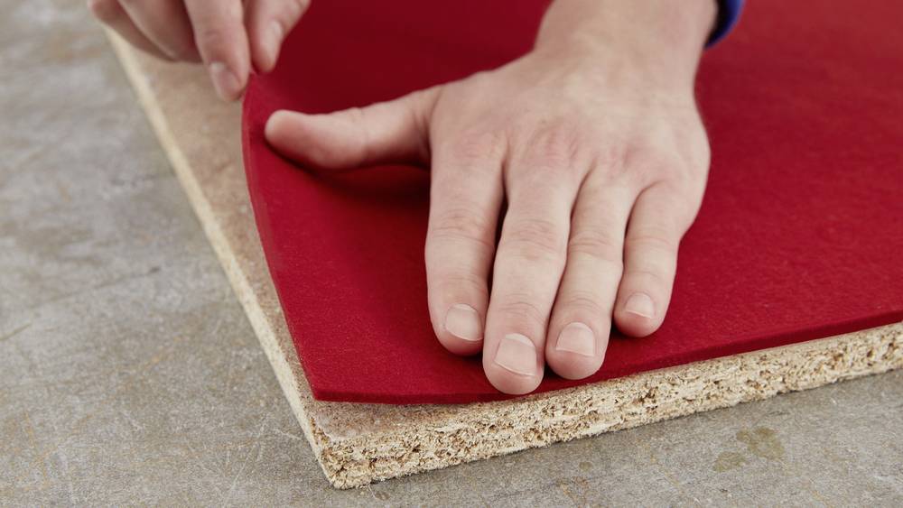 Eine Hand legt ein rotes Filzstück auf eine Spanplatte, vermutlich für eine Bastel- oder Bauarbeit.